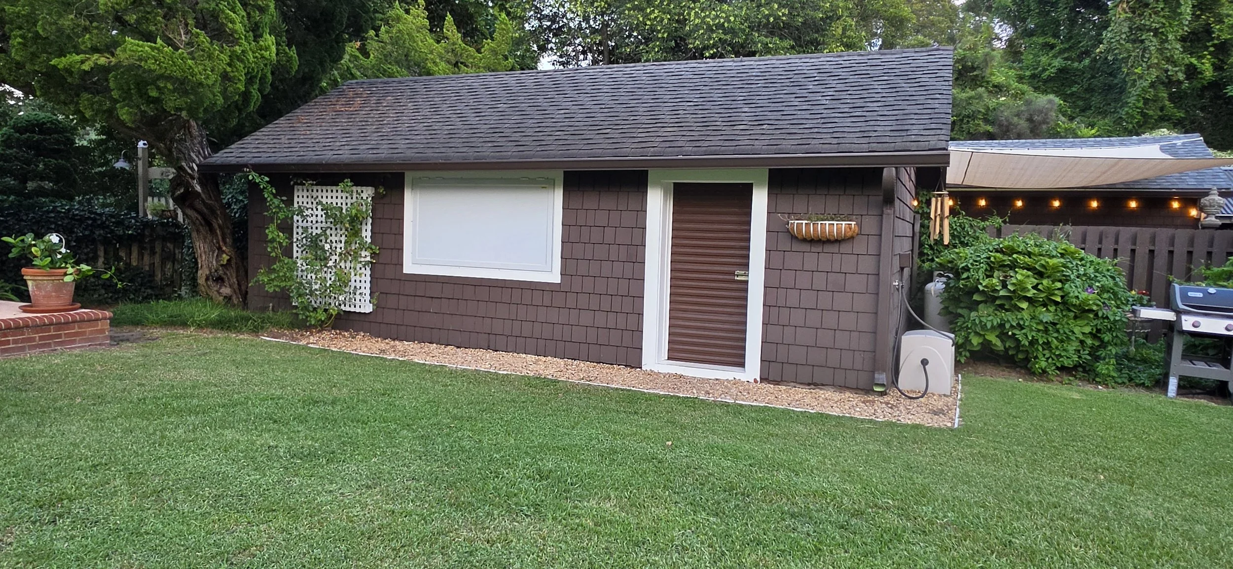 Backyard with a small brown shed, large green trees, a potted plant on a brick platform, and a barbecue grill. The shed has a window, a door with a brown shutter, and a hanging planter outside. String lights hang on the shed's side.