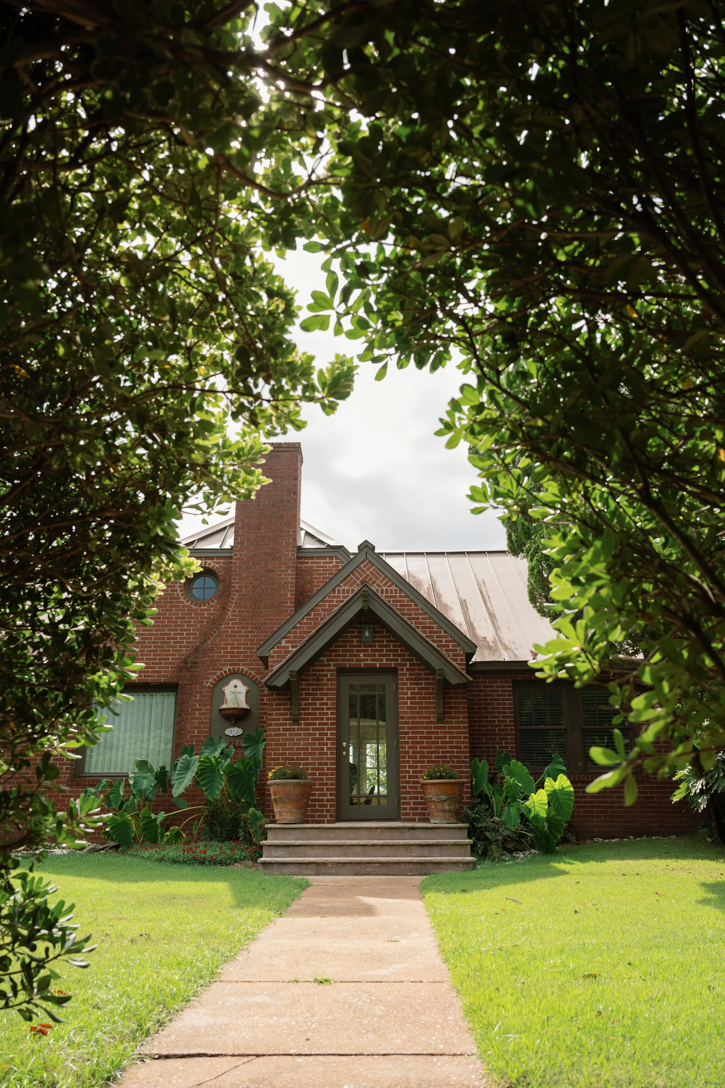 Red brick house with front steps, lush tropical landscaping, and a tree-lined walkway
