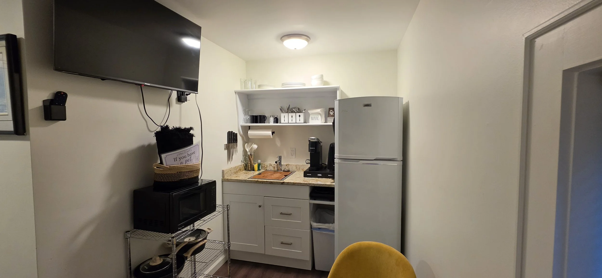 Kitchen with white cabinets, refrigerator, microwave, small sink, open shelf with plates, glasses, and utensils, coffee maker, wall-mounted TV, and a yellow chair in front.