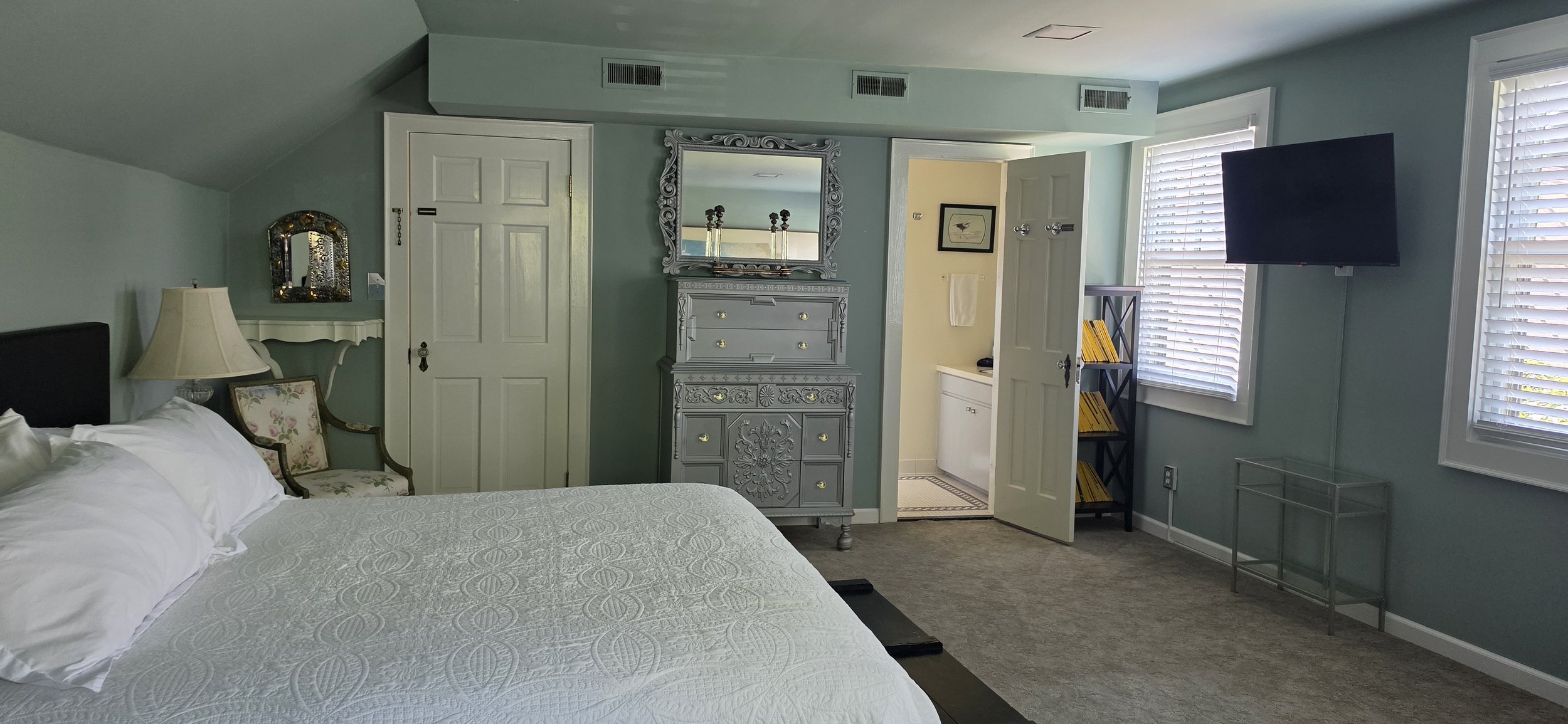 A cozy bedroom with light blue walls, a white bed with a quilt, a vintage armchair with floral upholstery, a bedside lamp, a decorative mirror above a small white table, a gray ornate dresser with a mirror, and a mounted flat-screen TV on the wall ne