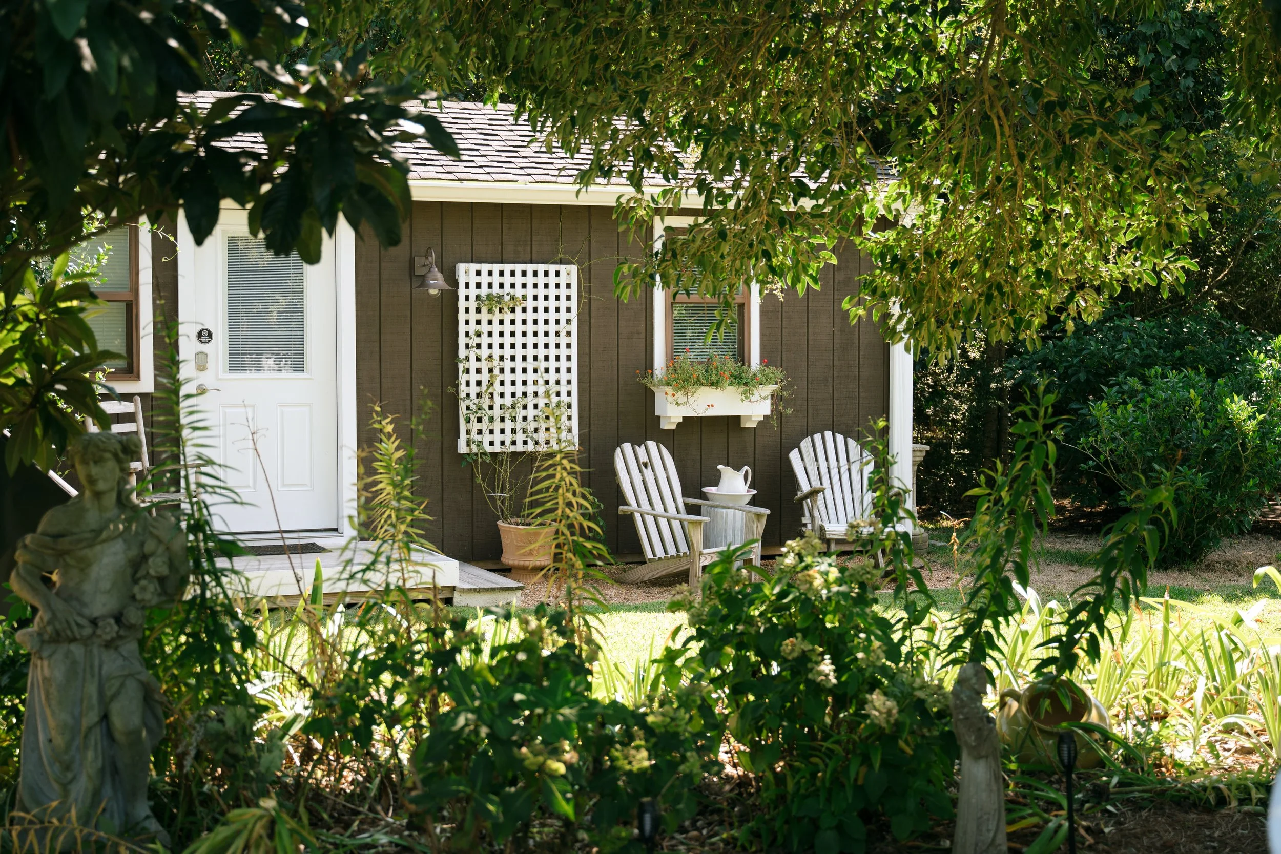 Cottage patio with white Adirondack chairs, trellis, planter box, and lush garden foliage