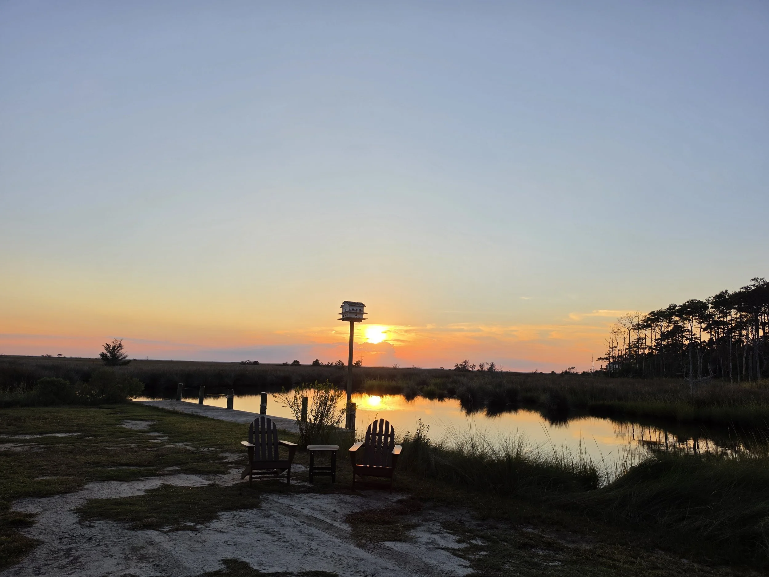 A sunset over a peaceful swamp with a small house on stilts in the distance, surrounded by trees and a small pond with two chairs facing the water.