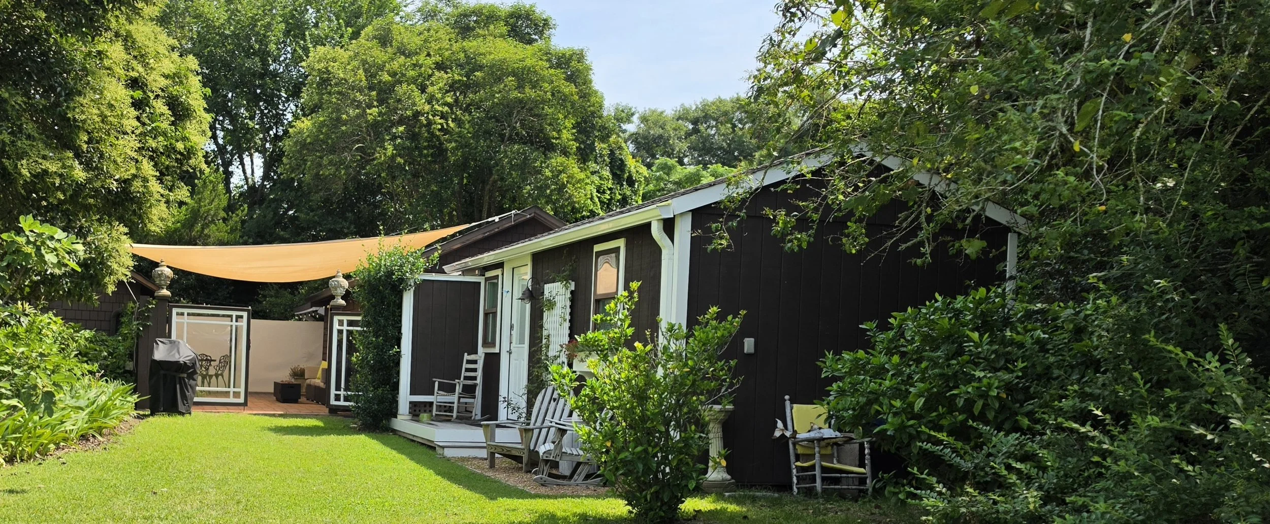 A backyard with green grass, lush trees, a dark brown house with white trim, a porch with chairs, an outdoor grill, and a shade sail over a patio area, all surrounded by landscaping.