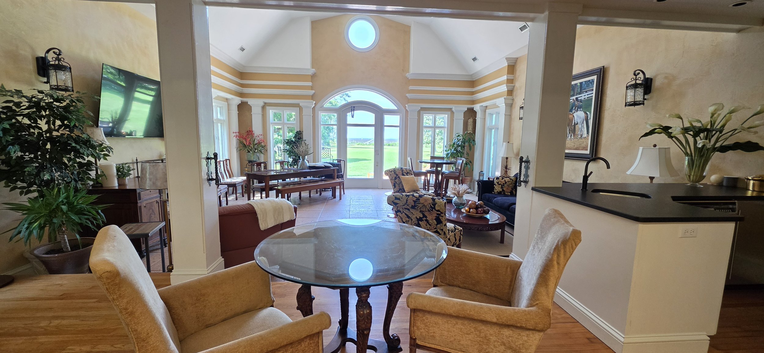 Bright living room with large windows and a view of a green landscape outside. The room has a mix of traditional and modern furniture, including a round glass dining table with beige armchairs, floral-patterned armchairs, and a dining table with chairs near the windows. Decor includes plants, framed artwork, vases with flowers, a large wall-mounted TV, and a small kitchen area with a black countertop and a plant in a vase.