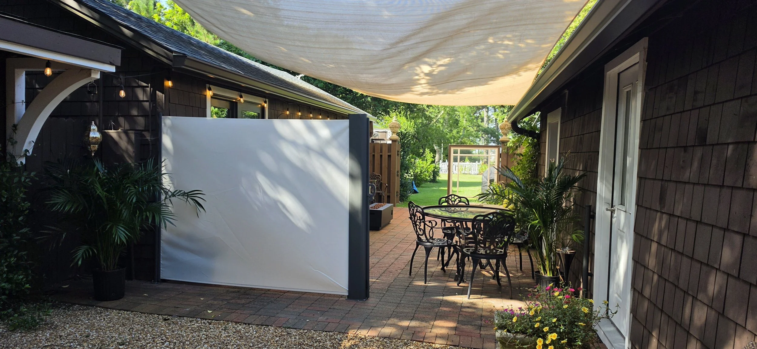 A cozy backyard patio with a round metal table and four chairs, potted plants, a white privacy screen, string lights, a shade sail overhead, and a view of a lush green lawn with trees and a white picket fence in the background.
