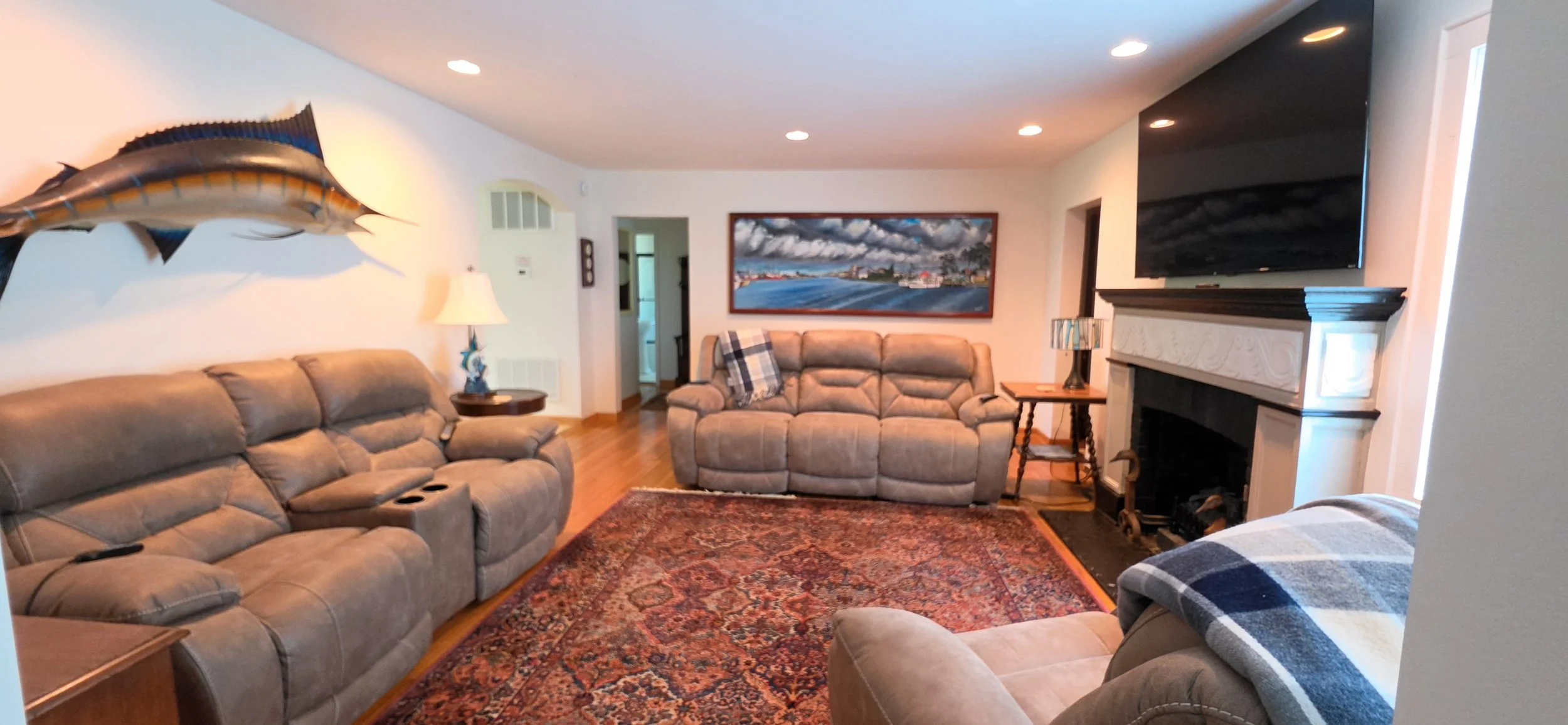 Living room with beige sofas, a patterned rug, a framed painting of the outdoors, a fireplace, and a wall-mounted TV.