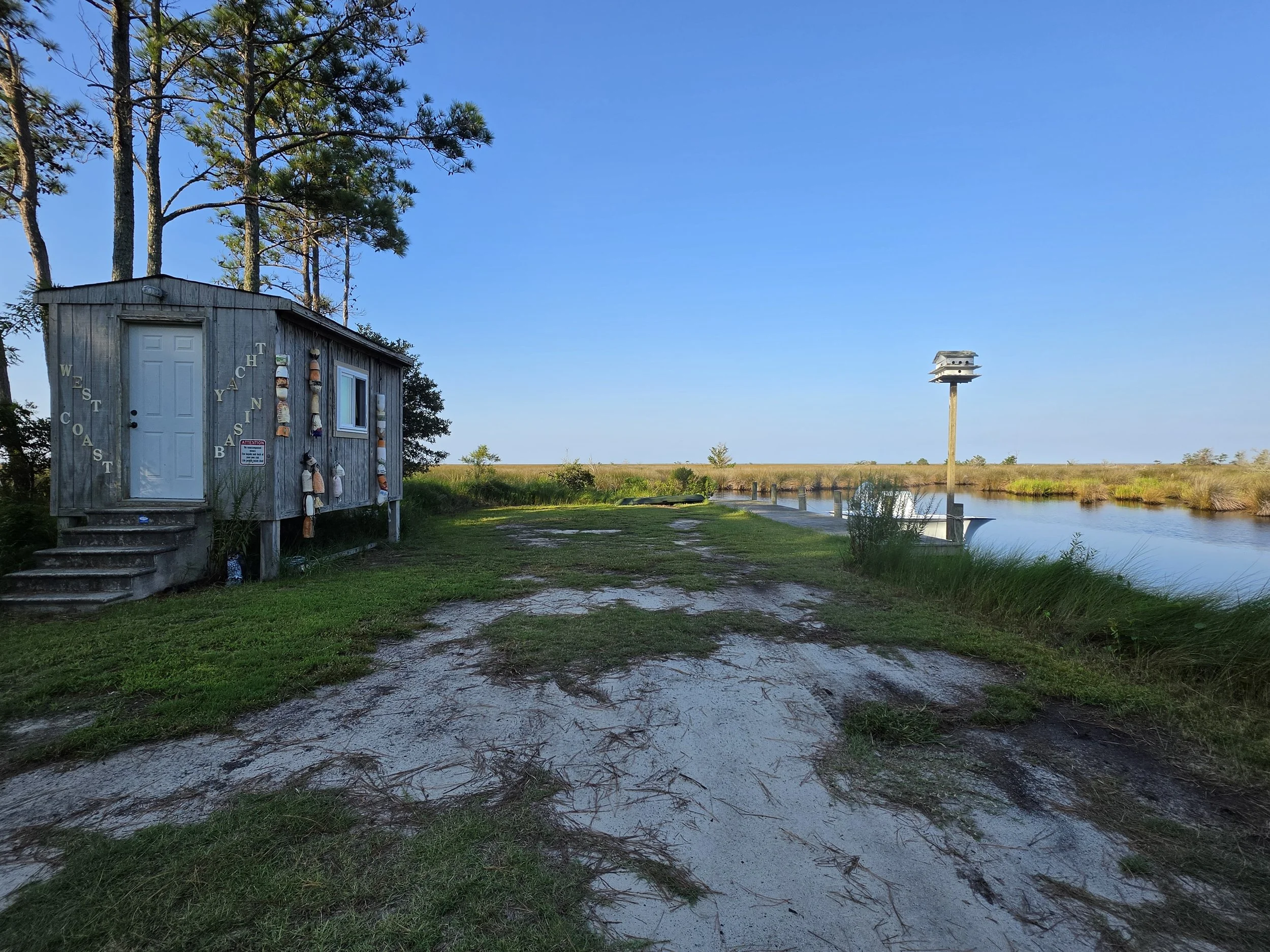 A small wooden cabin situated near a body of water with a birdhouse on a tall pole and tall pine trees in the background under a clear blue sky.