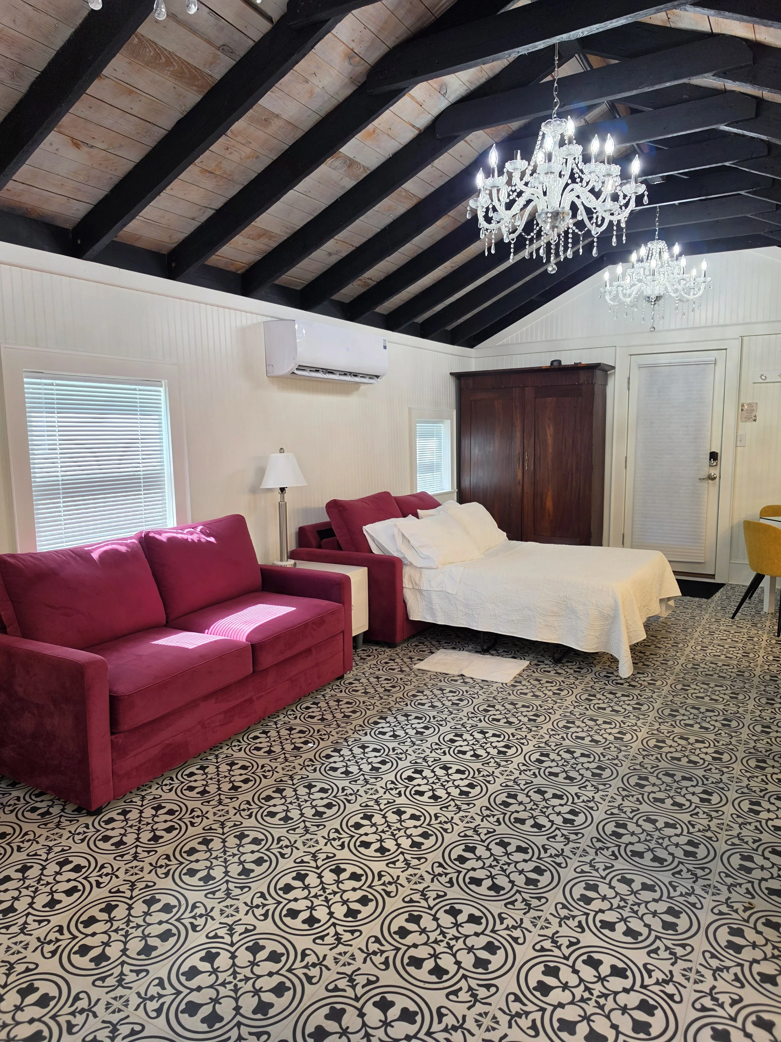 A bedroom with a patterned black and white tile floor, a white bed with a white bedspread, a red velvet couch, a wooden armoire, two chandeliers hanging from the ceiling, and two windows with blinds.