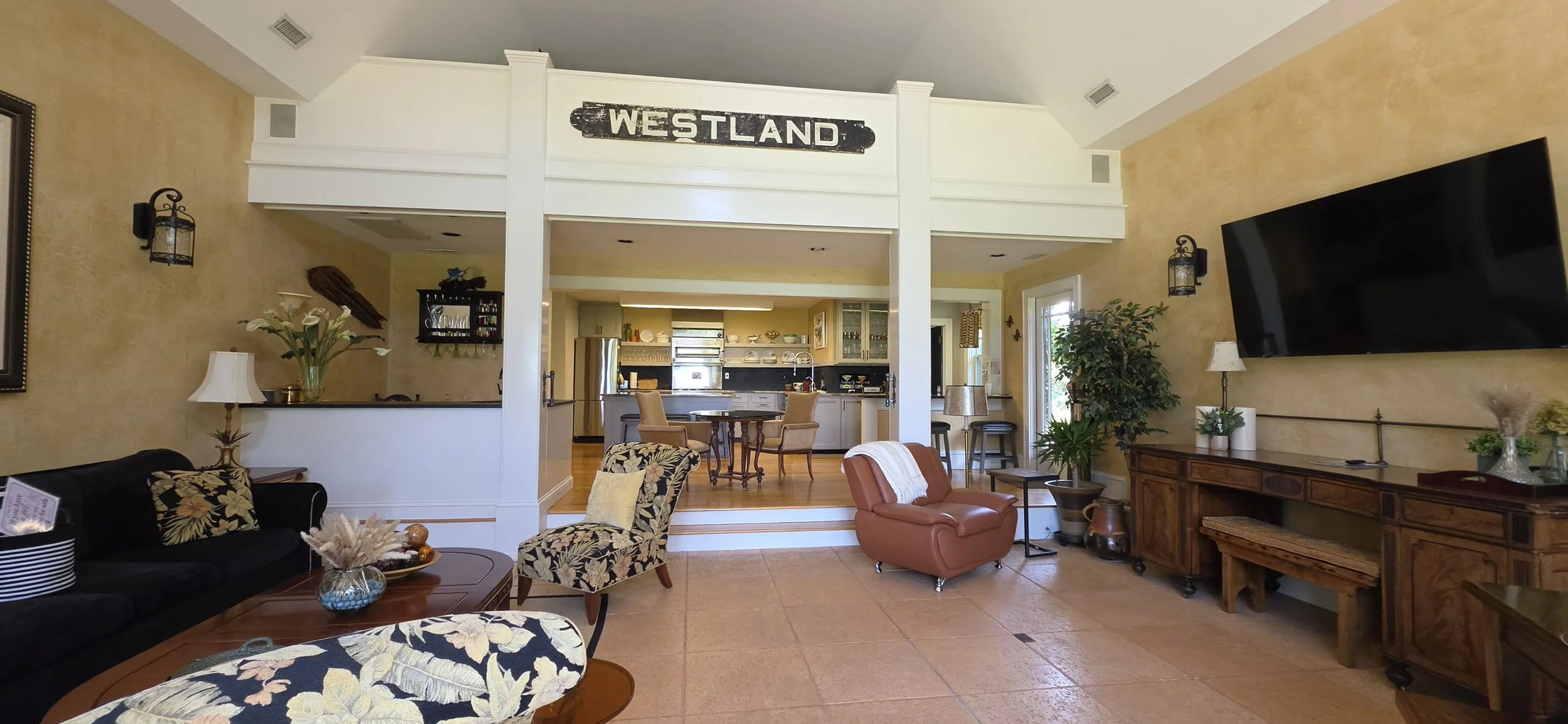Living room with black sofa, floral armchair, and brown leather chair; coffee table with decorative items; large wall-mounted TV; trees and plants; open kitchen and dining area in background; sign labeled 'Westland' above doorway.