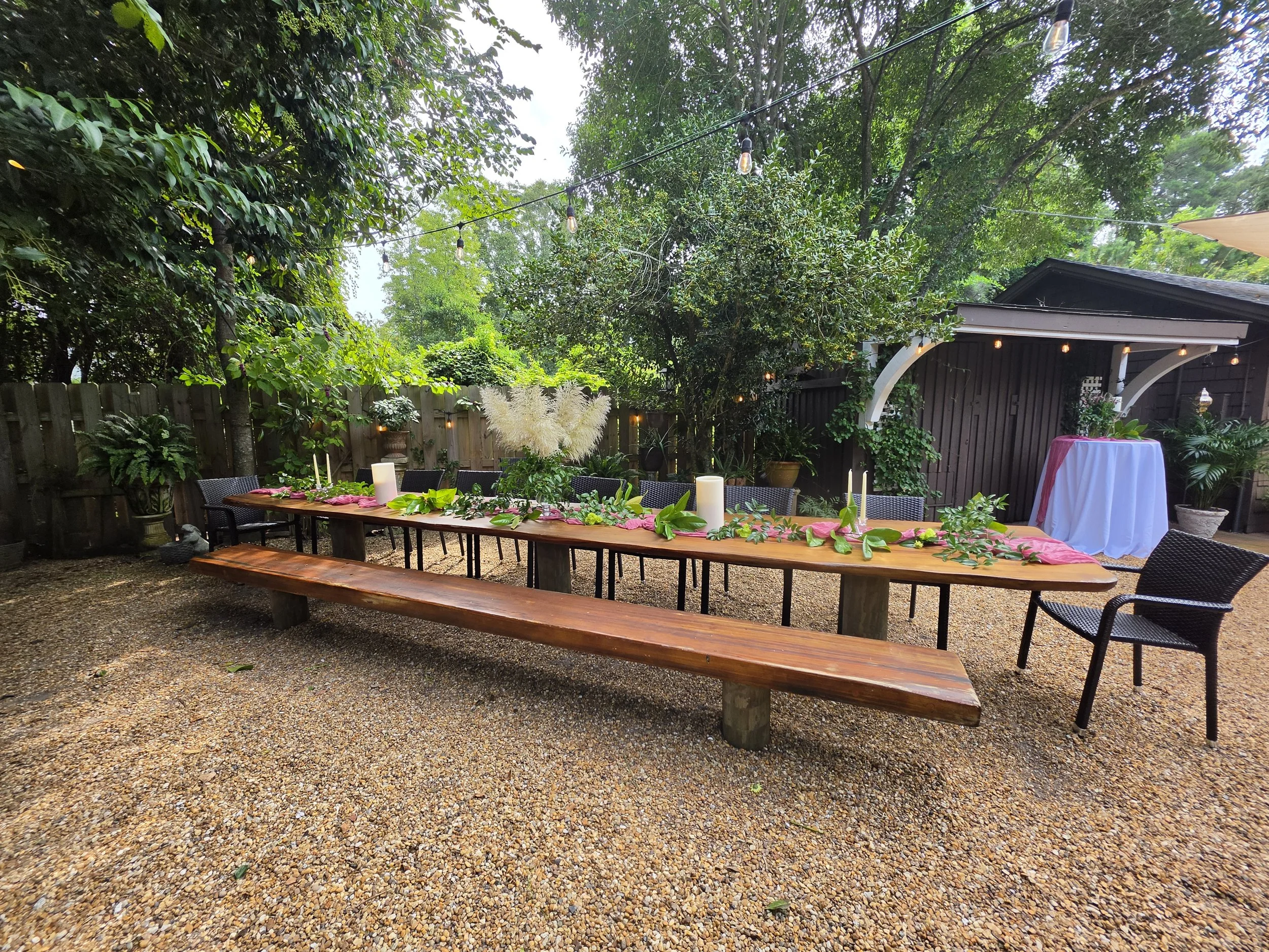 An outdoor dining area with a wooden table and black chairs decorated with a pink table runner and greenery, surrounded by trees with string lights overhead and a small wooden shed or pavilion to the right.