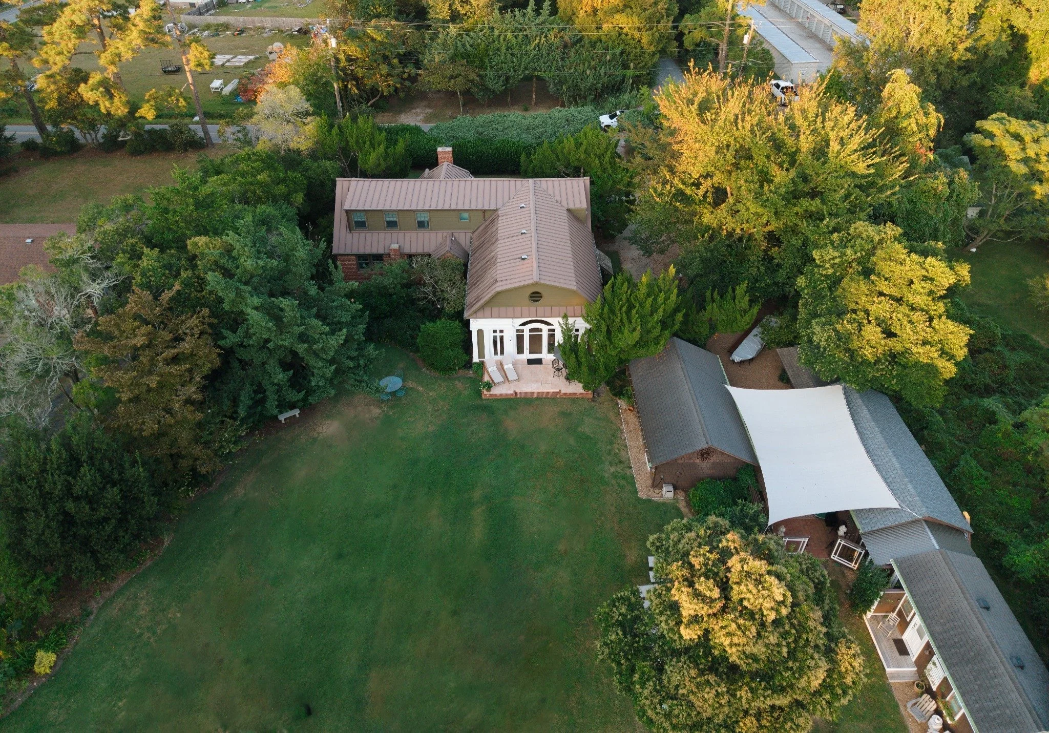 An aerial view of a residential backyard with a house, a well-maintained lawn, and various trees and bushes. The house has a tan exterior with a metal roof and a small front porch. The backyard includes a patio with outdoor furniture and is surrounded by tall trees and dense greenery. Neighboring properties with additional houses and yards are visible, along with utility poles and wires.