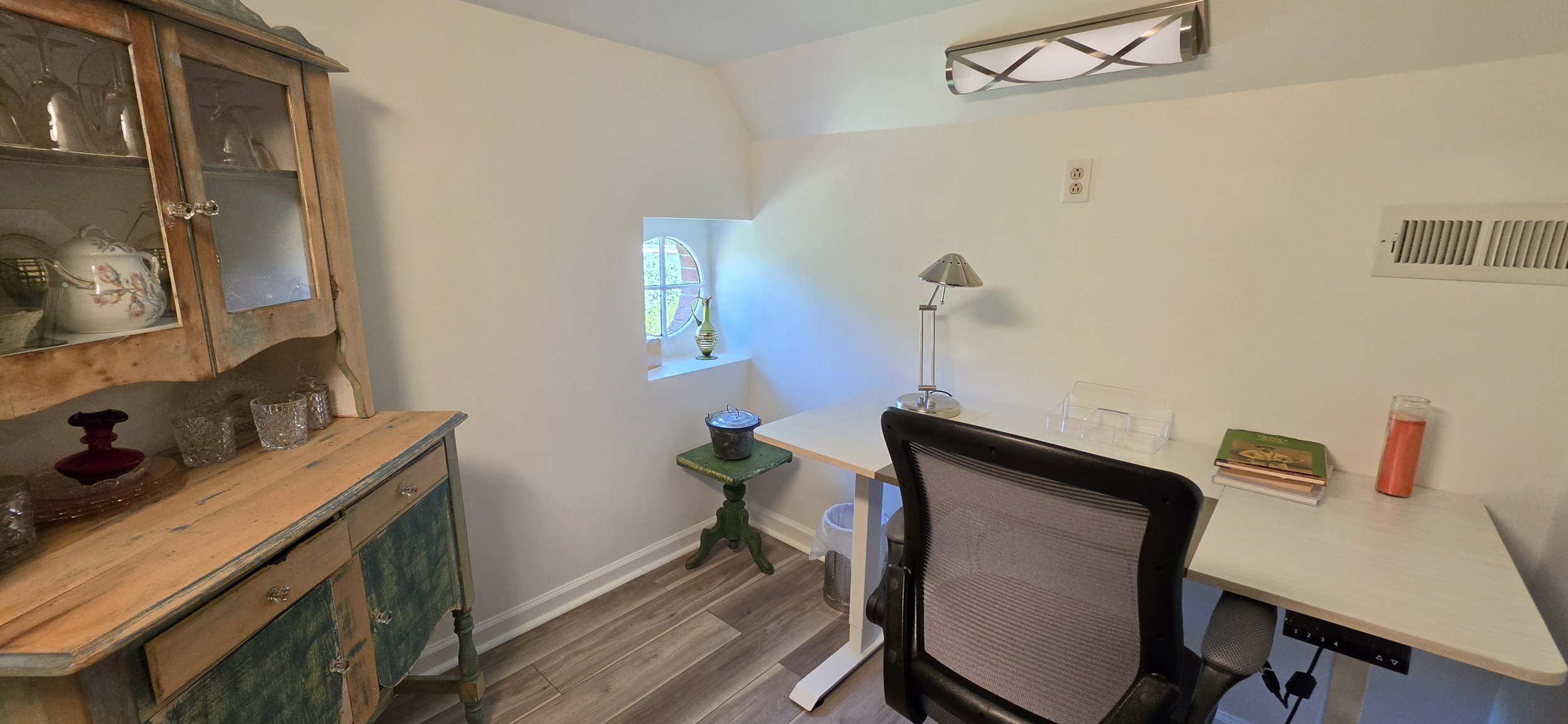 A small home office with a white desk, black office chair, and a window with a decorative wooden frame. There is a vintage wooden china cabinet on the left with glass doors displaying china and glassware. On the desk, there are books, a lamp, a candl