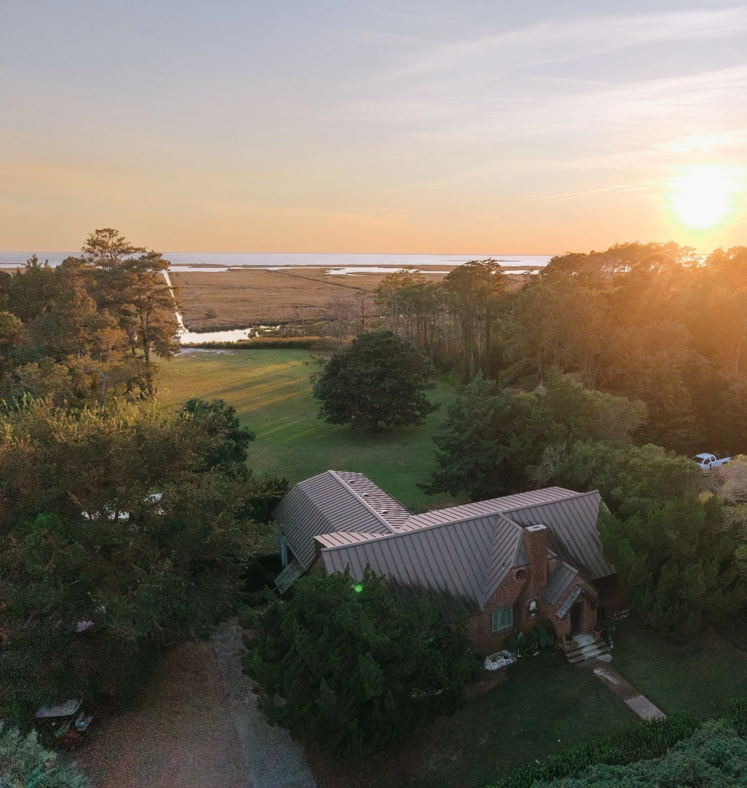 A sunset view over a coastal landscape with trees, a grassy area, a house with a metal roof, and a body of water in the distance.