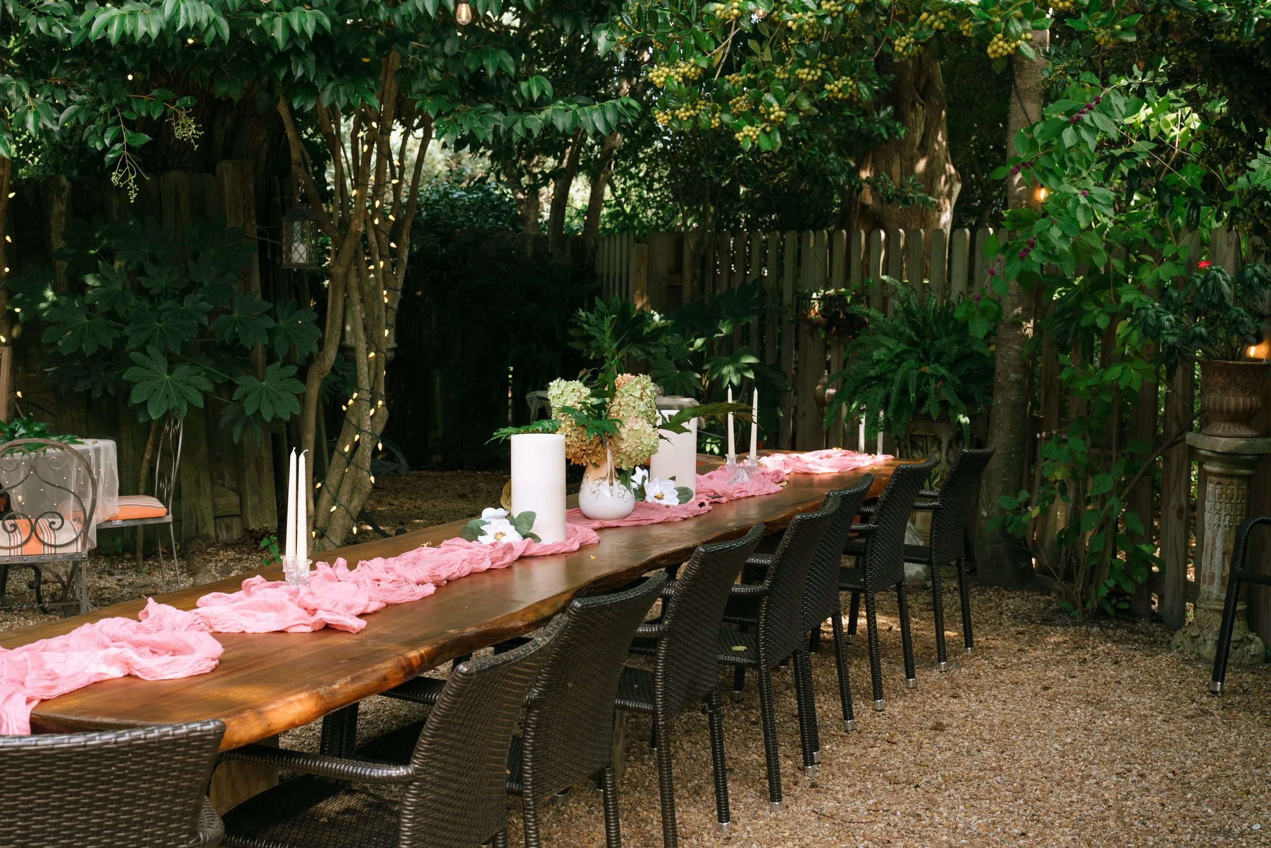Outdoor garden dining table decorated with pink table runners, candles, flowers, and wicker chairs beneath lush greenery
