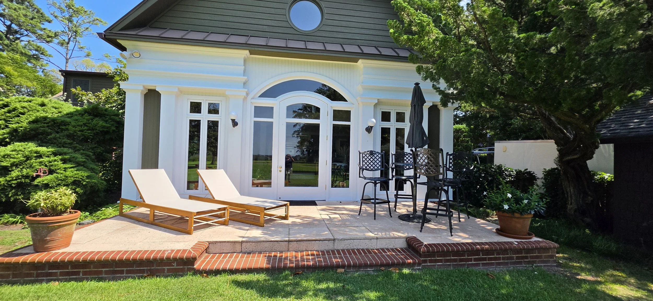 A backyard patio area in front of a house with large glass doors, two lounge chairs, potted plants, and a table with chairs shaded by an umbrella, surrounded by greenery and trees.