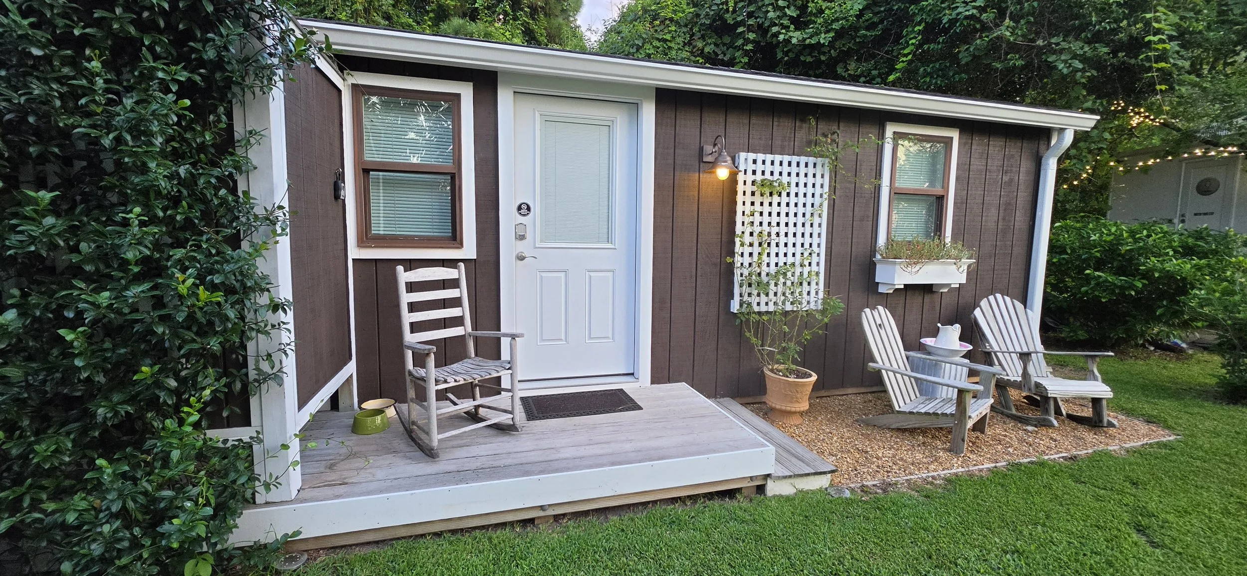 The image shows the front of a small brown house with a white door and two windows. There is a small porch with two wooden chairs, one with a small green pet bowl nearby. To the right, there is a potted plant and a white wall-mounted planter with plants. A white watering can is on one of the chairs. A wall-mounted light is turned on, illuminating part of the porch. The background shows trees and bushes, creating a lush, green setting.