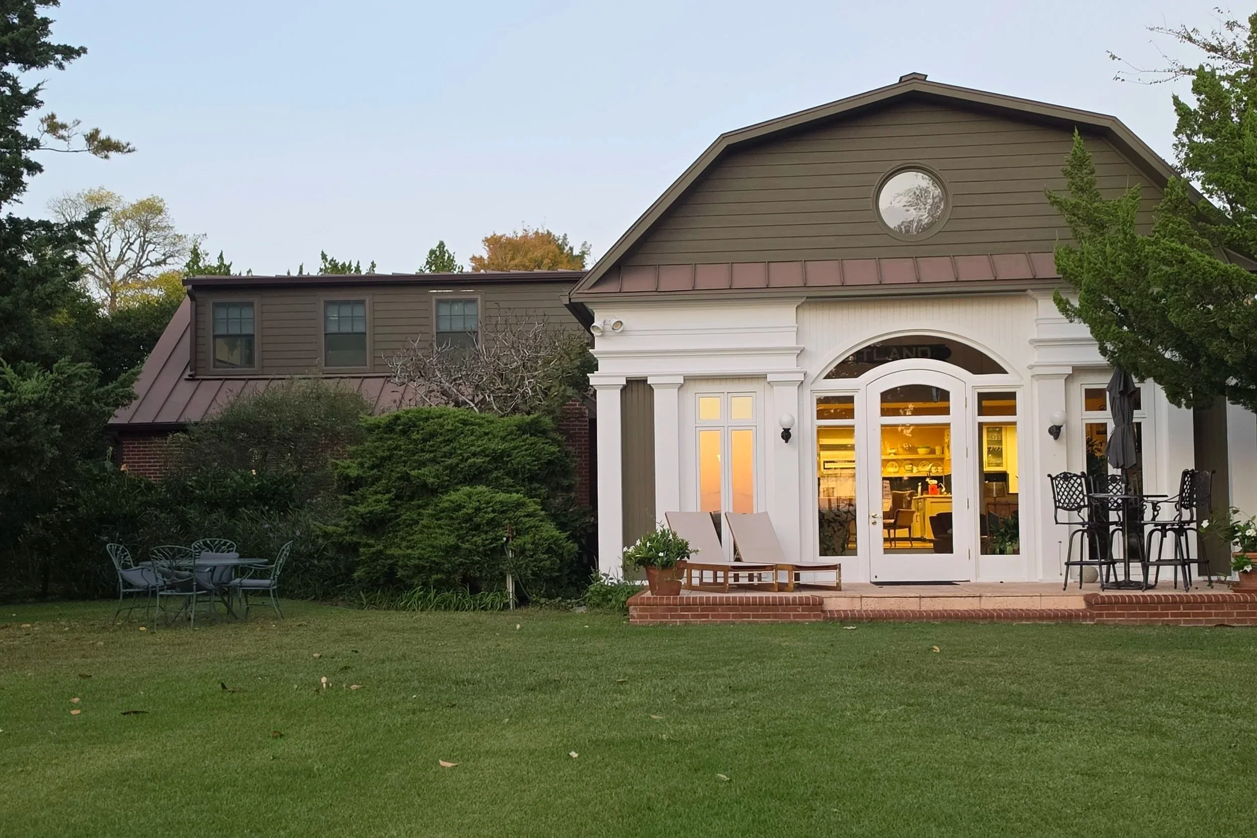 A house with a backyard, illuminated indoor lights visible through large glass doors, surrounded by green grass and trees, with outdoor furniture on a patio.