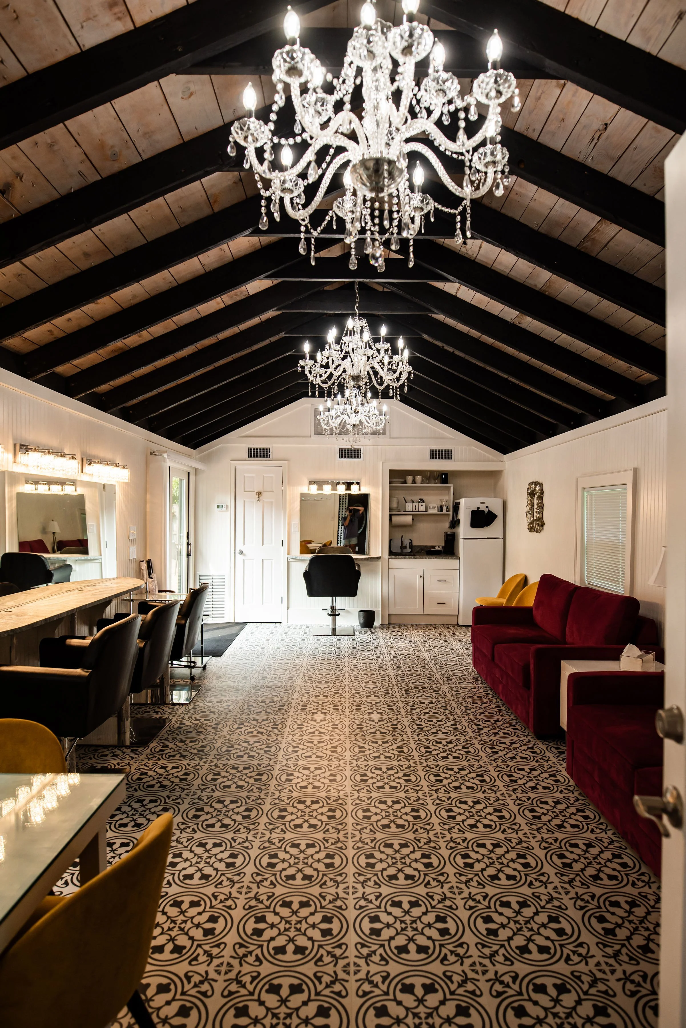 Elegant salon interior with crystal chandeliers, exposed wood beams, patterned tile flooring, a red sofa, bar seating, and kitchenette