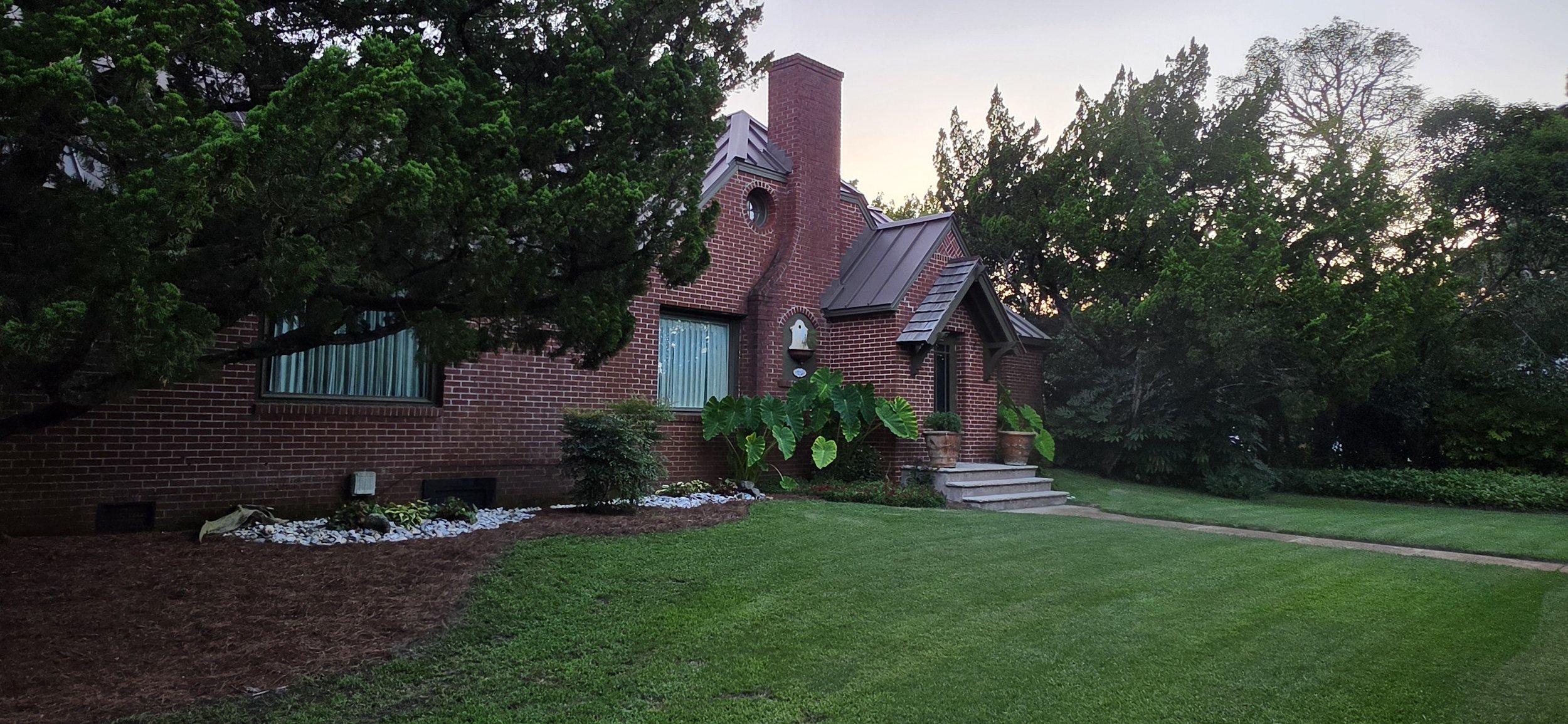 A red brick house with a metal roof, chimney, and steps leading up to the front door in a well-maintained yard with green grass and large trees surrounding the house at dusk.