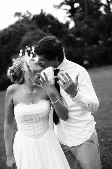 A black-and-white photo of a bride and groom sharing a kiss outdoors, with trees in the background. The bride is in a strapless wedding dress with a pearl necklace, and the groom is in a dress shirt with a tie.