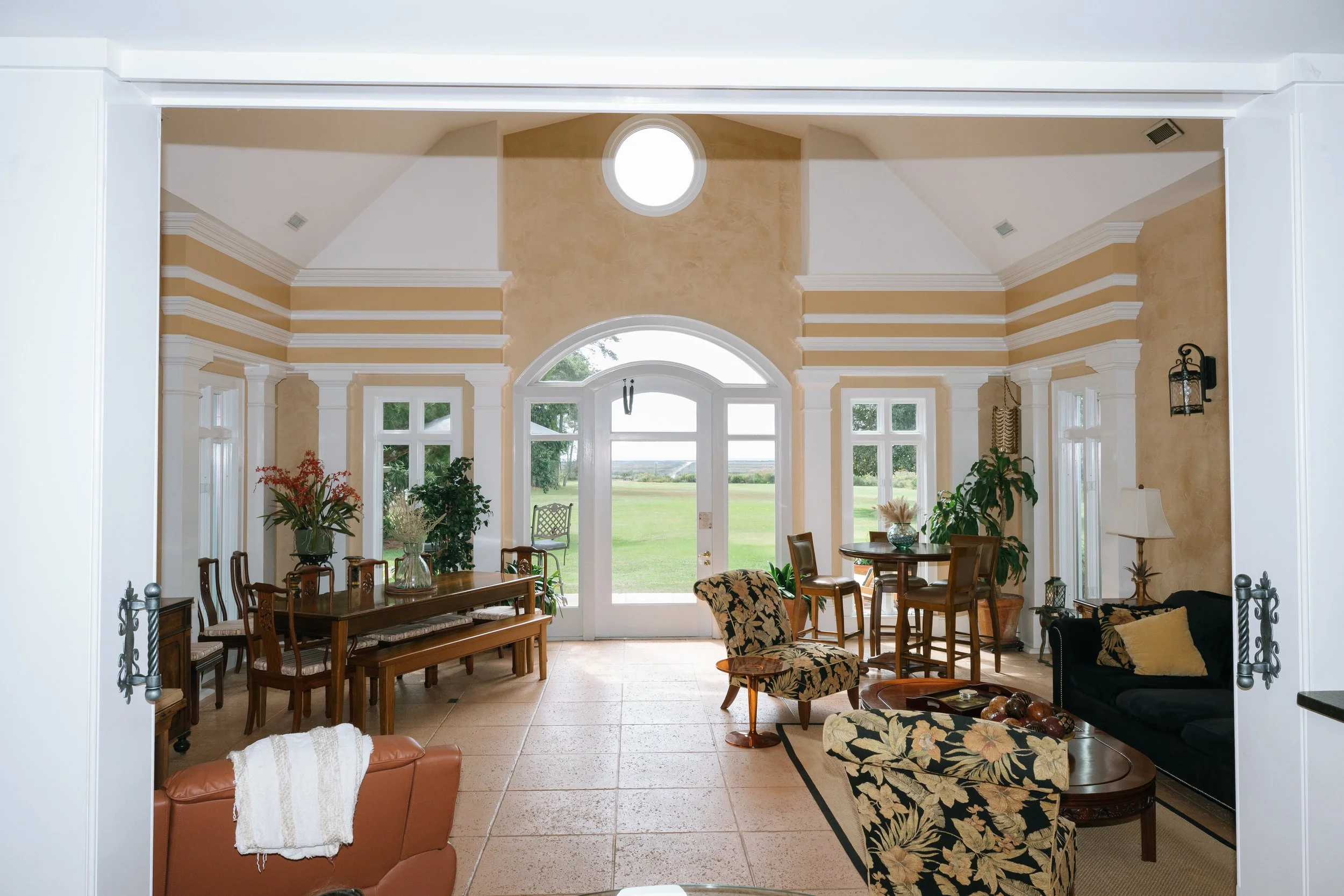 Elegant sunroom with ornate white molding, large arched windows, traditional furniture, plants, and views of a green lawn