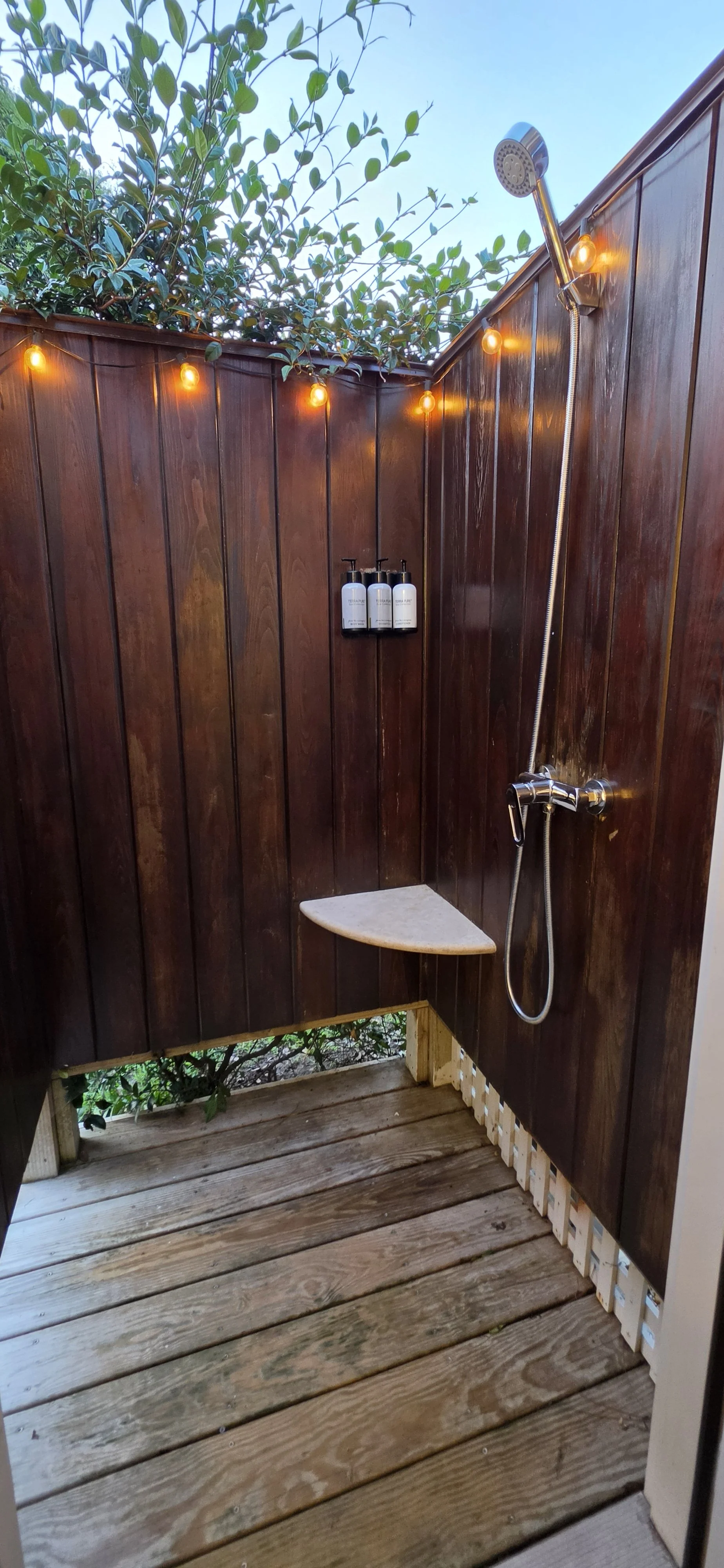 Outdoor wooden shower area with string lights, a small shelf with soap bottles, a corner seat, and greenery visible at the top.