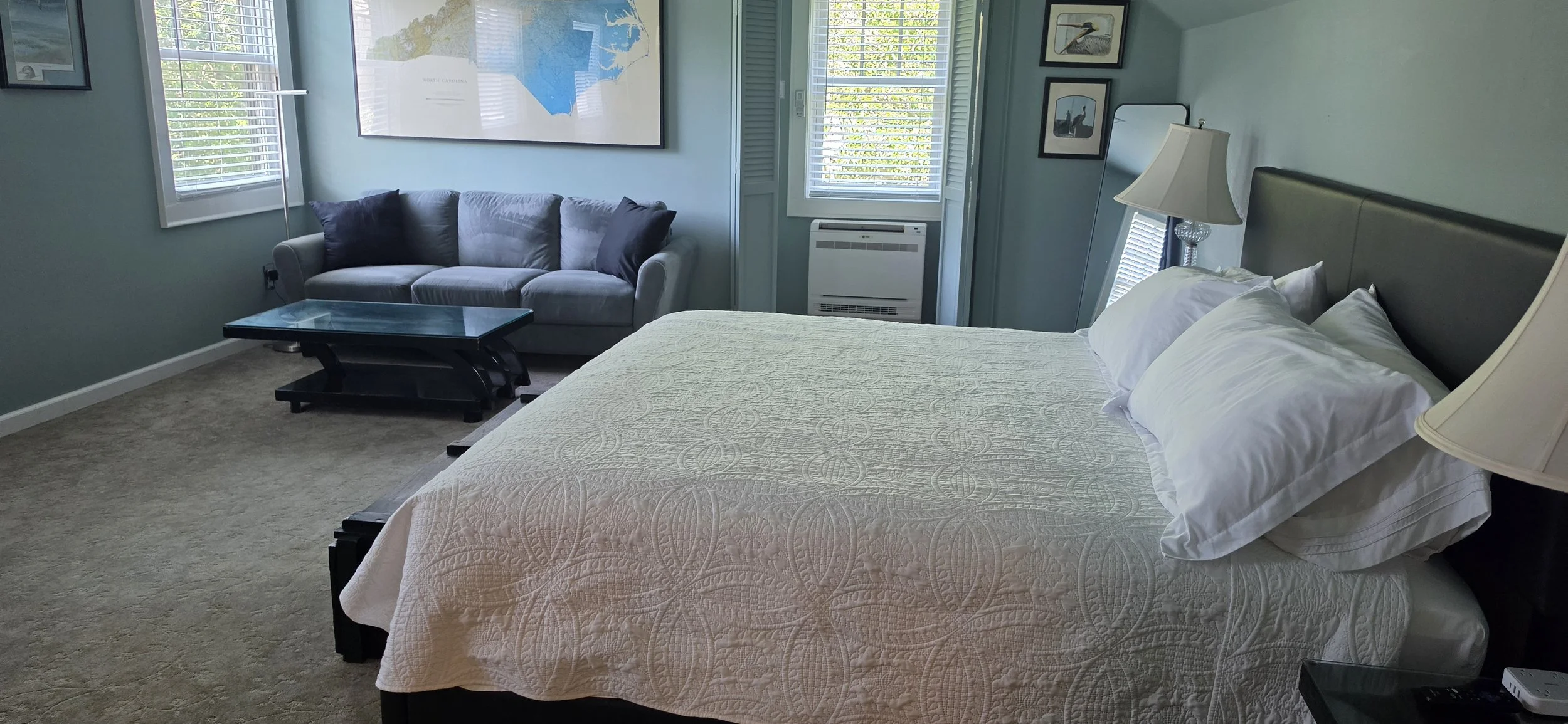 Bright bedroom with a king-size bed, white quilt, gray loveseat, black coffee table, lamps, windows, and framed artwork.