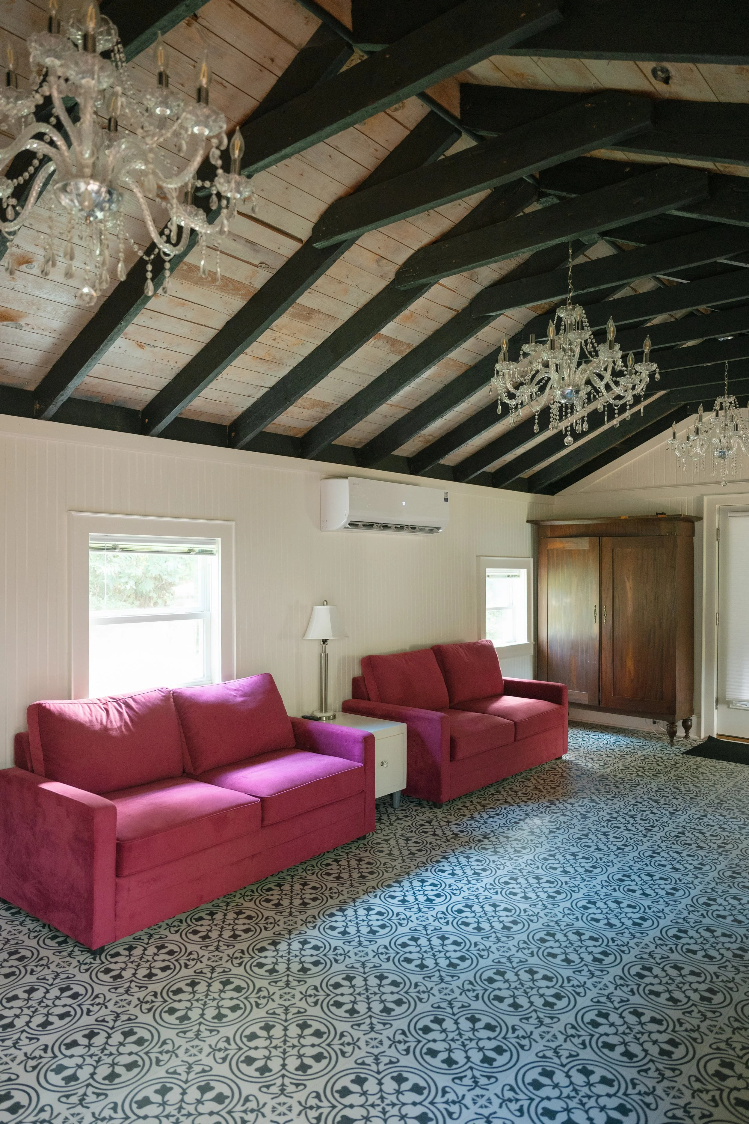 Bright living room with pink sofas, patterned carpet, crystal chandeliers, exposed dark ceiling beams, and a wooden cabinet.