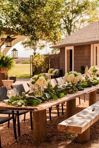Outdoor dining table decorated with white flowers, greenery, and candles, set for a meal on a sunny day.