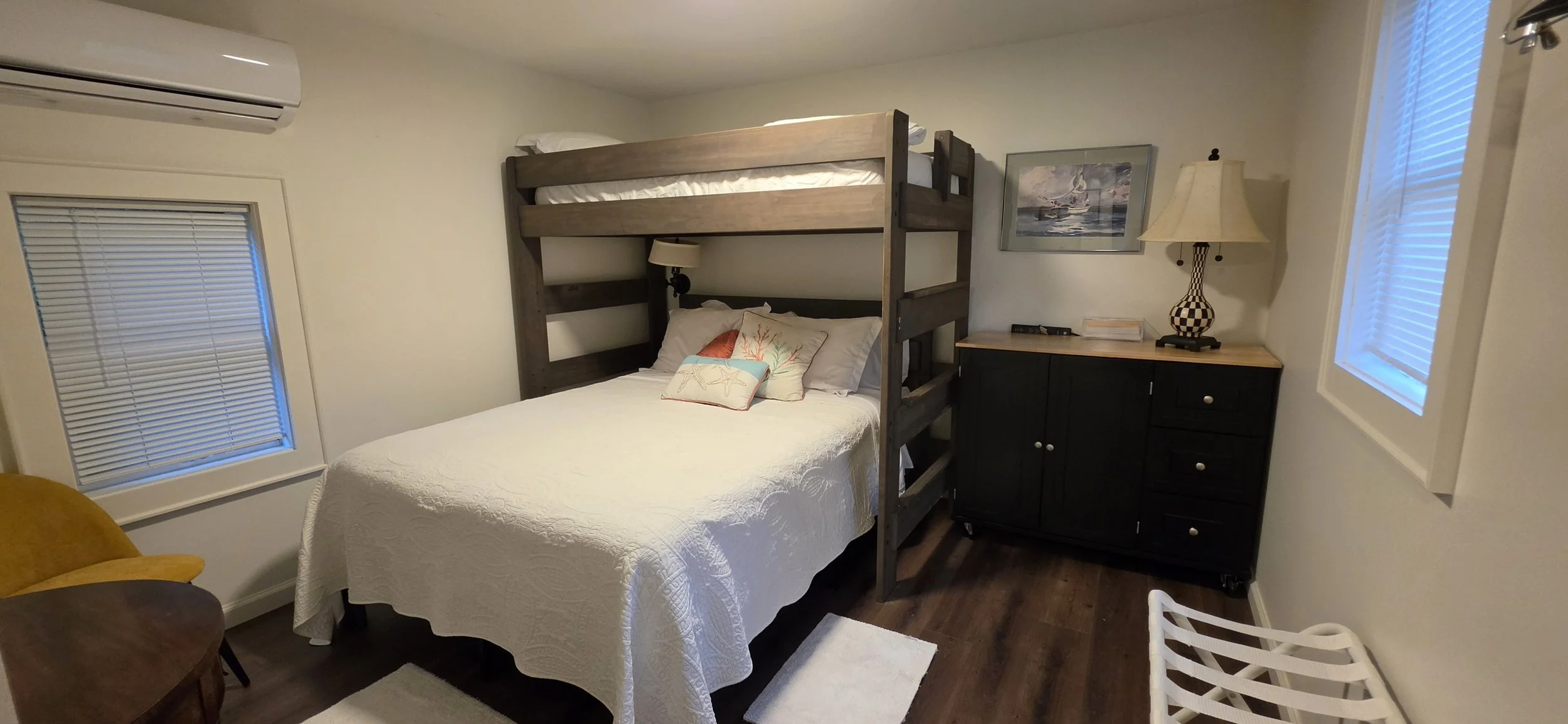 A bedroom with two windows, a bed with a white quilt, pillows, and a wooden bunk bed frame. There is a dark wood dresser with a lamp, books, and a picture on the wall.