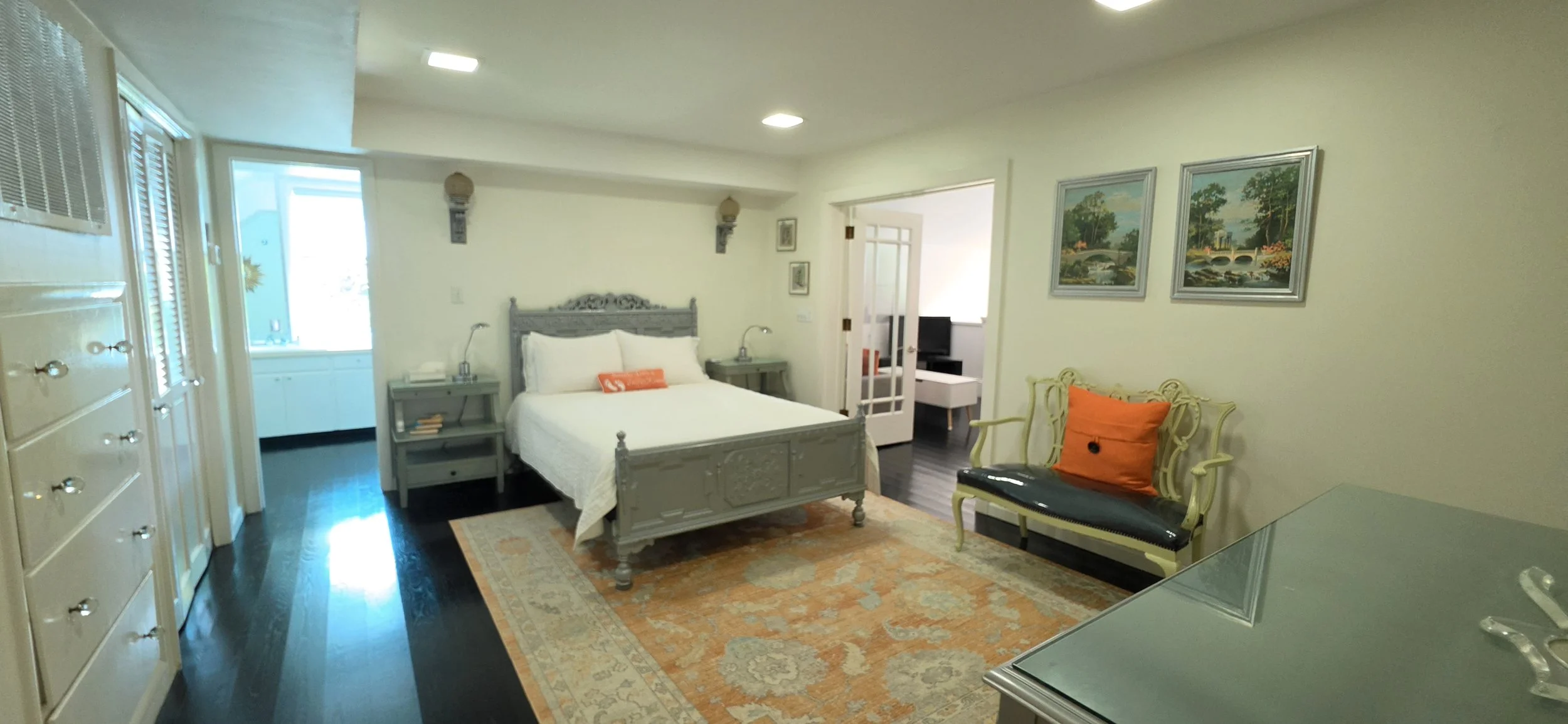 A bedroom with a large bed, two nightstands, a bench with an orange cushion, and two paintings of bridges on the wall.