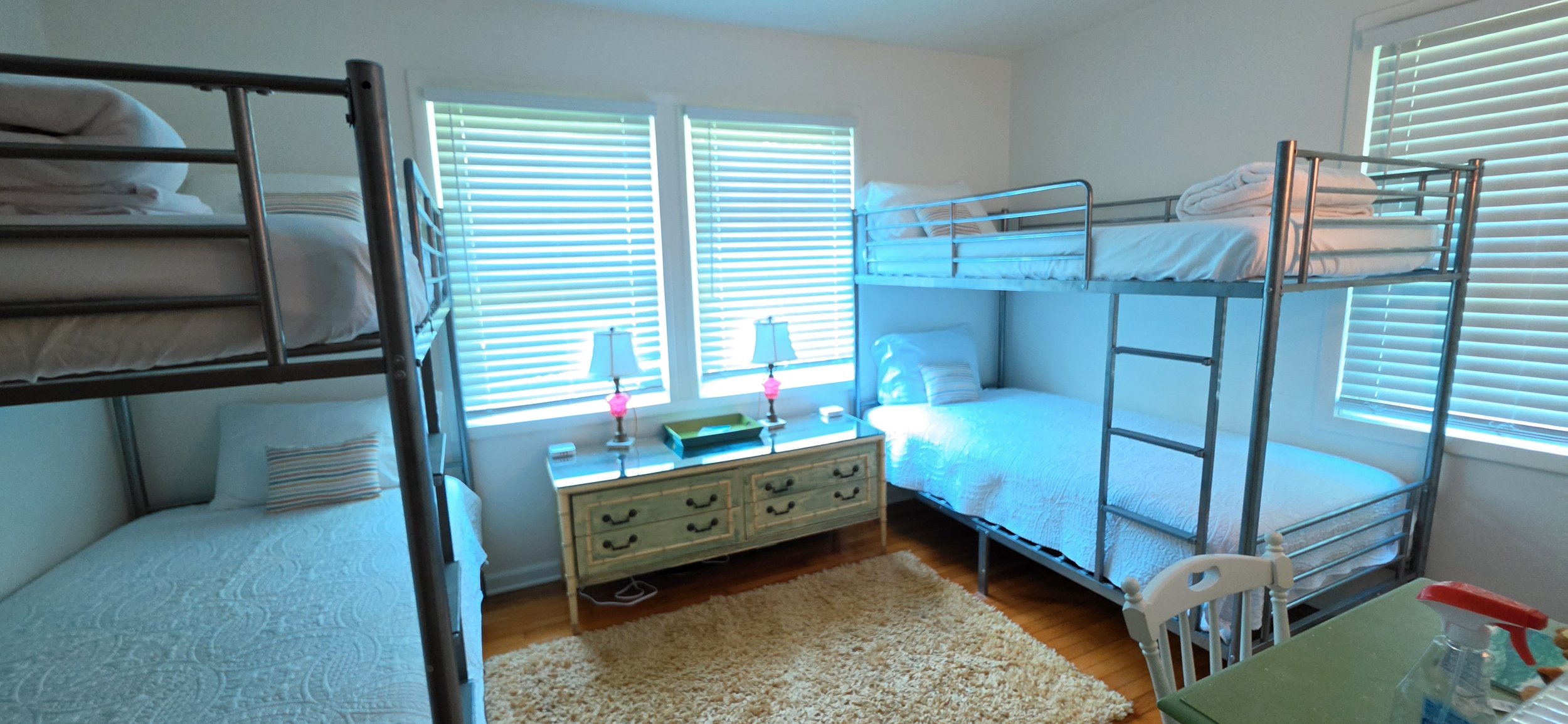 A bedroom with three beds, including two metal bunk beds and one single bed. There are three windows with blinds, a dresser between the windows with lamps on top, and a yellow rug on a wooden floor. A small chair and table are partially visible in th