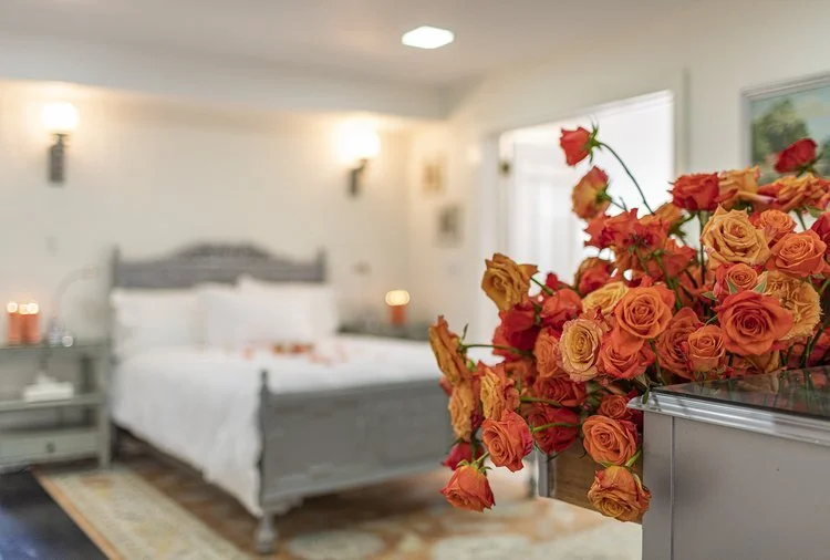 Orange roses in the foreground of a guest bedroom at Bostwick’s Magnolia Manor, with a gray bed frame, white bedding, and soft lighting.