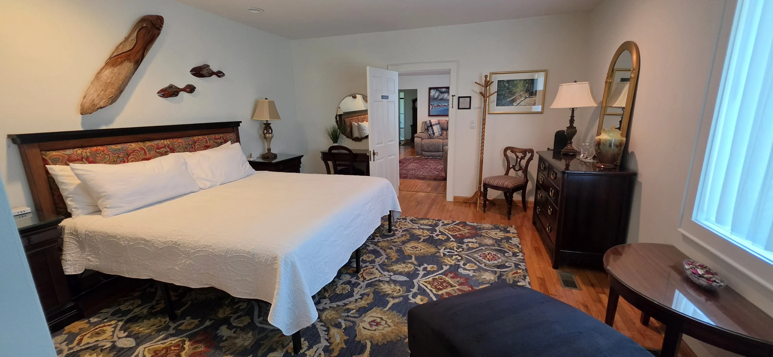 A bedroom with a bed covered in a white quilt, a patterned headboard, a colorful rug, a dresser with a mirror, and various wall decorations including a wooden fish art piece. There is a window with blinds, and a small seating area visible through the