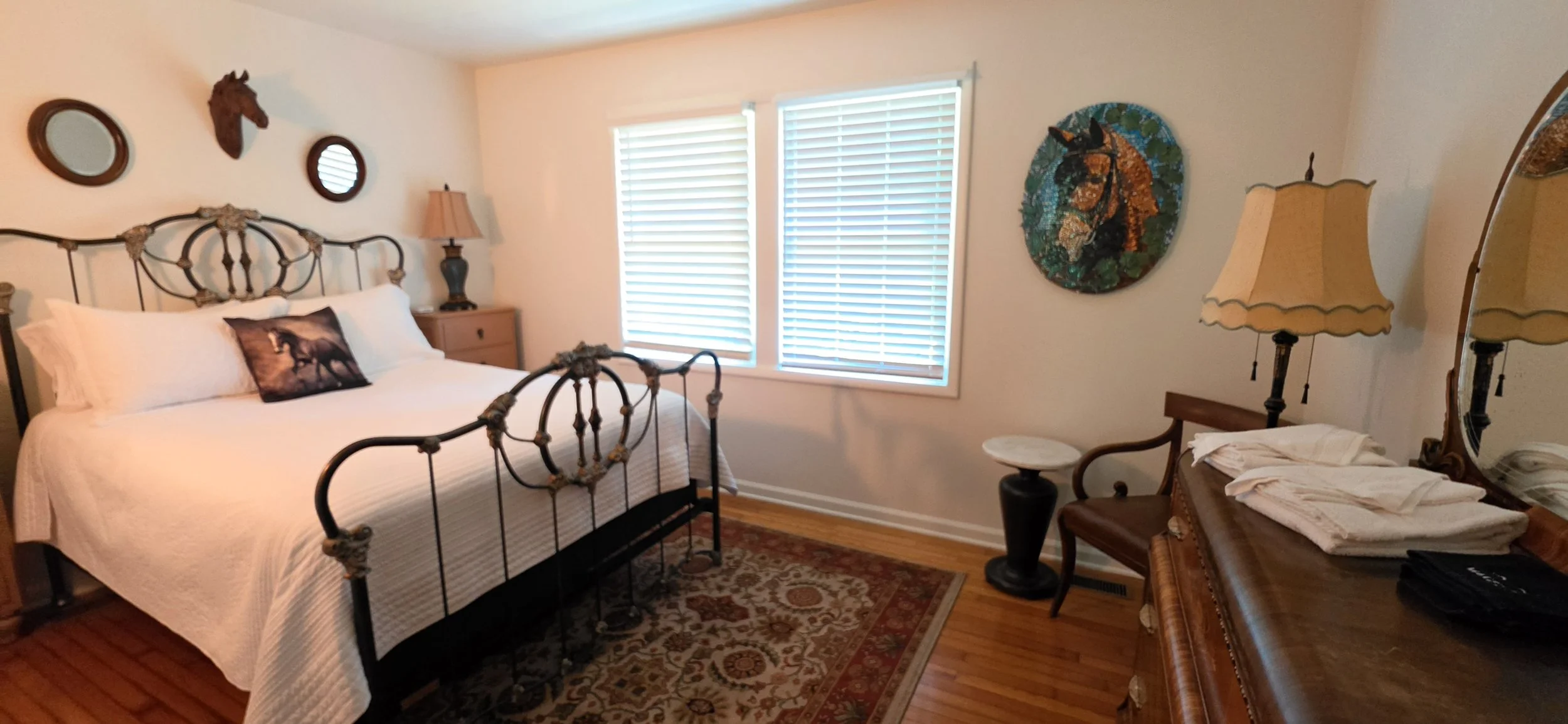 A bedroom with a metal bed frame, white bedding, and a decorative pillow featuring a horse. There are two nightstands with lamps, one with a horse head wall decor above it. The room has two windows with blinds, a decorative tapestry of a horse on the