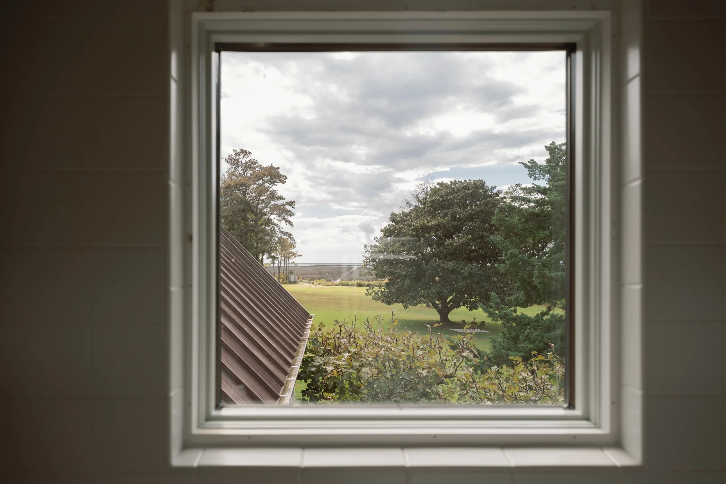 View through a window of a green lawn, trees, waterfront marsh, and cloudy sky