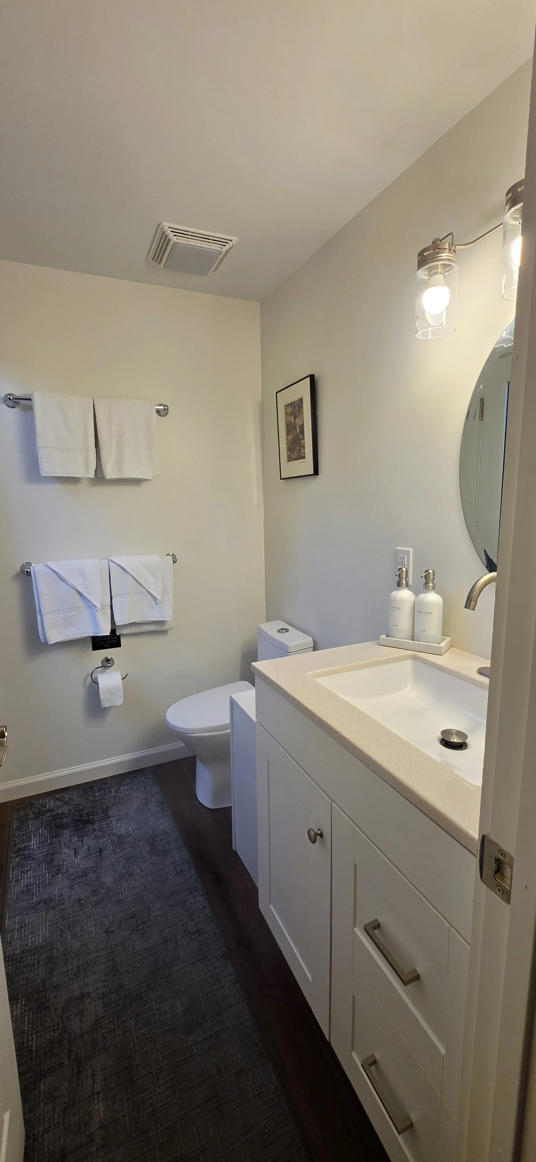 A clean bathroom with a white sink vanity, a toilet, two white towels on a silver towel rack, a small framed picture on the wall, a round mirror, a light fixture with two exposed light bulbs, and a dark textured rug on the floor.