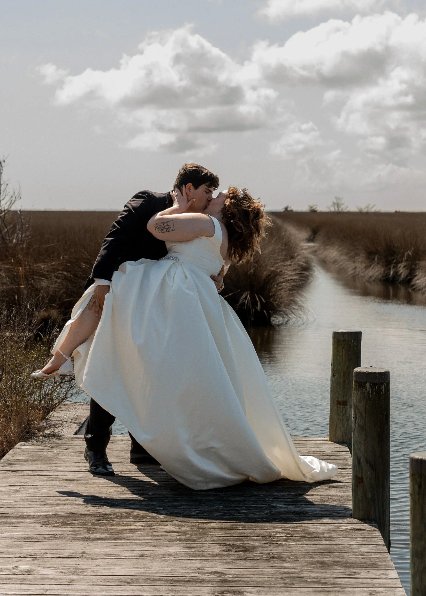 A couple dressed in formal wedding attire shares a kiss on a wooden dock beside a body of water, with a cloudy sky overhead.