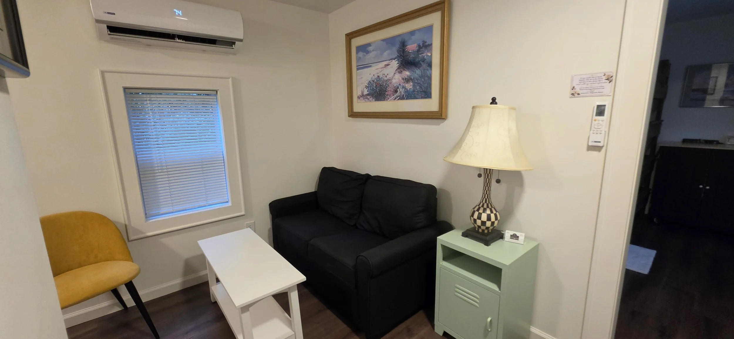 A small living room with a yellow chair, a black sofa, a white small table, a green cabinet, a table lamp with a checkered base, a framed painting of a beach scene on the wall, a window with blinds, and an air conditioning unit mounted above the wind