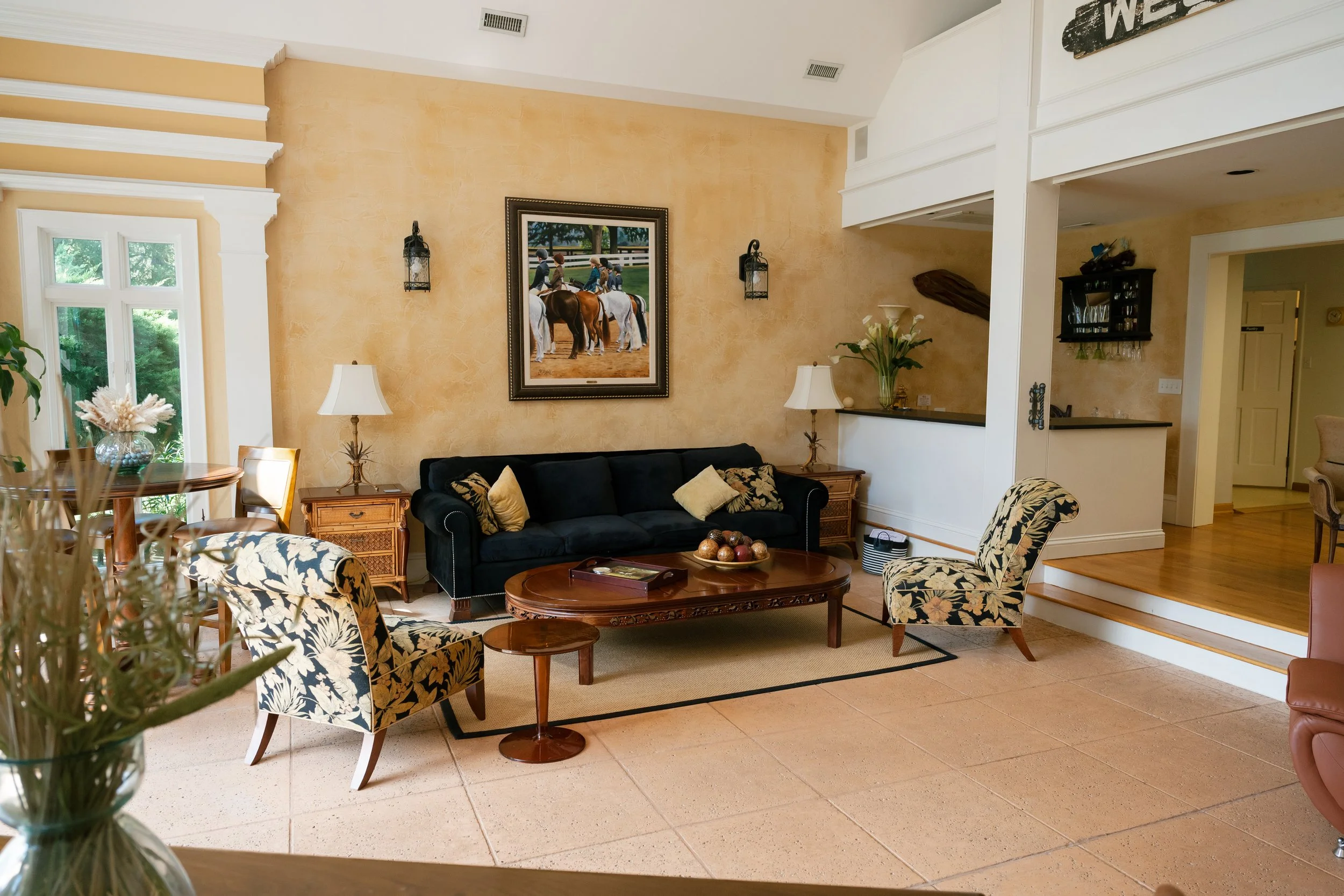 Elegant living room with navy sofa, floral armchairs, carved coffee table, framed equestrian artwork, and adjacent home bar