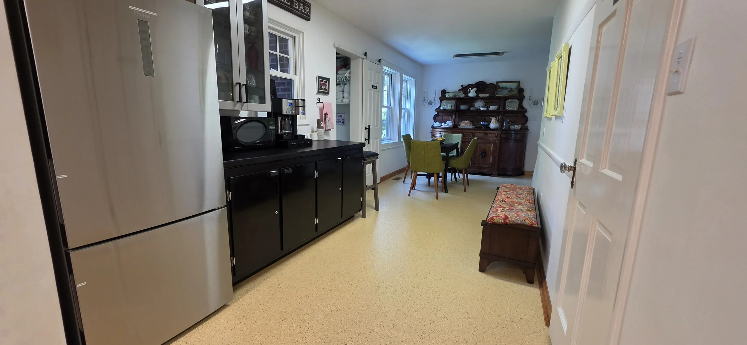 The image shows a cozy dining area with a wooden table and four green chairs. There is a dark wooden hutch with decorative dishes on it against the back wall. To the left, a partial view of a modern kitchen countertop with a black cabinet and a stain