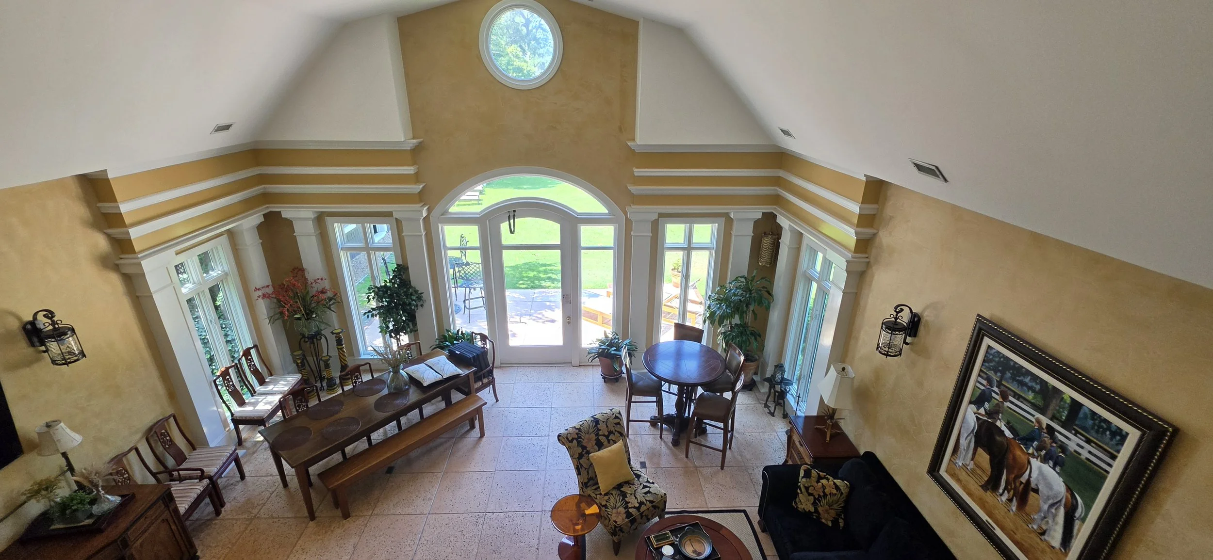 Living room with large windows and glass door leading outside, yellow walls, ceiling with a circular window, furniture including a dining table, chairs, and decorative plants.