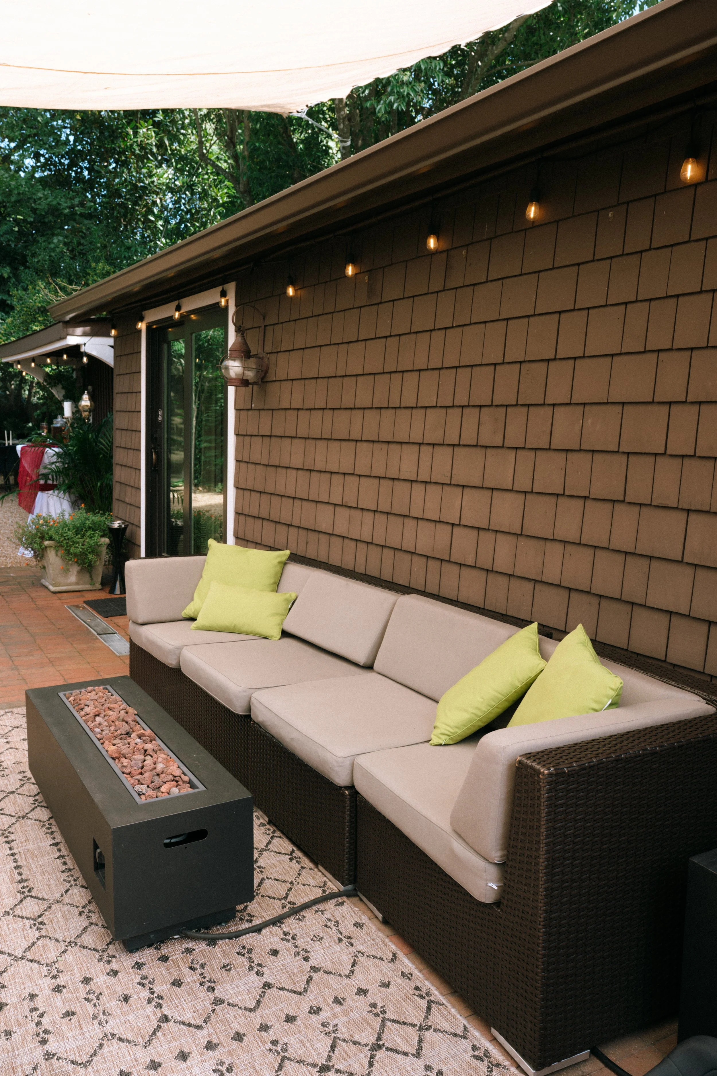 Covered outdoor patio with wicker sectional seating, lime-green pillows, string lights, and a rectangular fire pit beside a shingled wall.