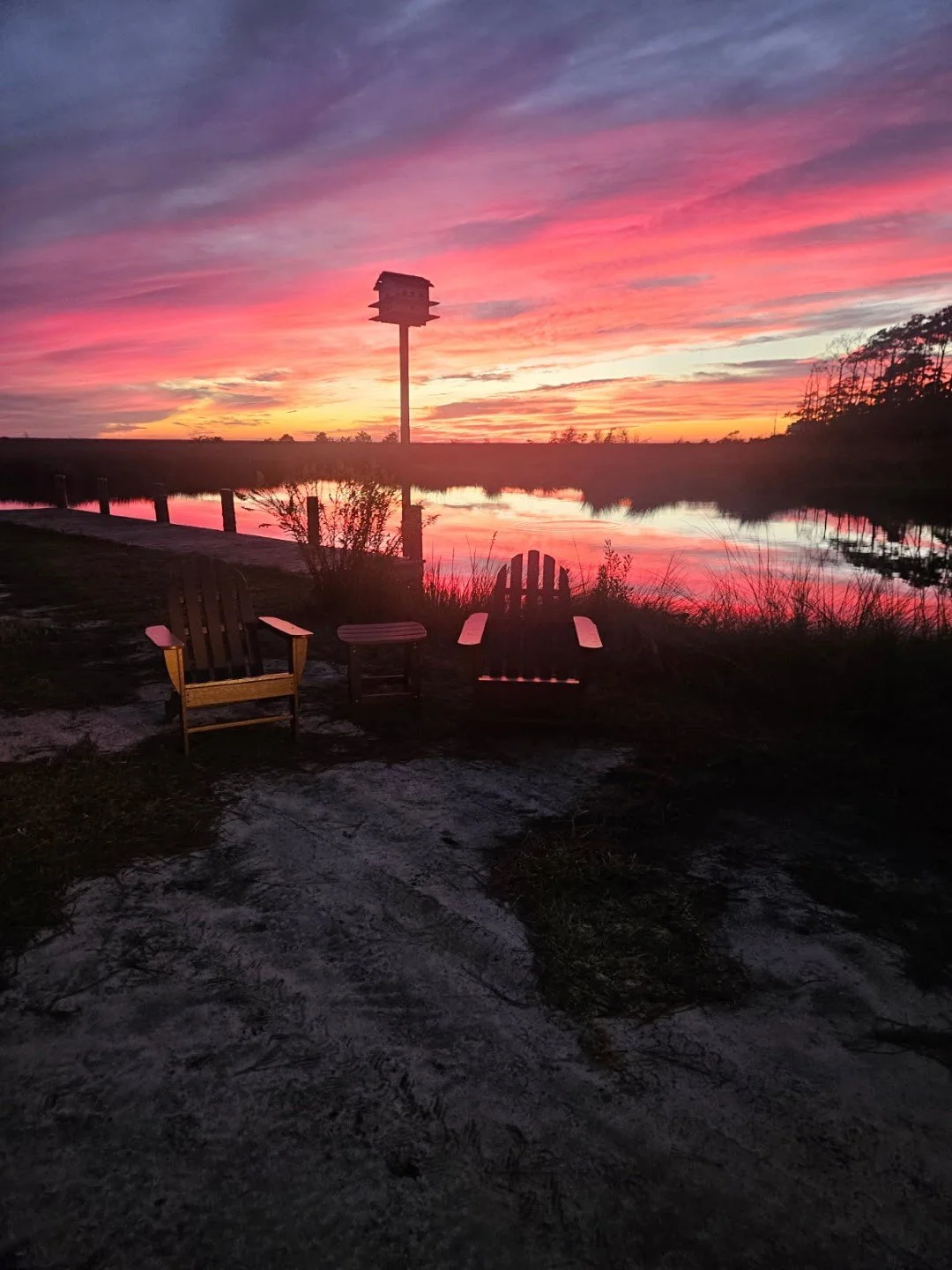Adirondack chairs beside a calm lake reflecting a vibrant pink and purple sunset, with a birdhouse silhouetted in the background.