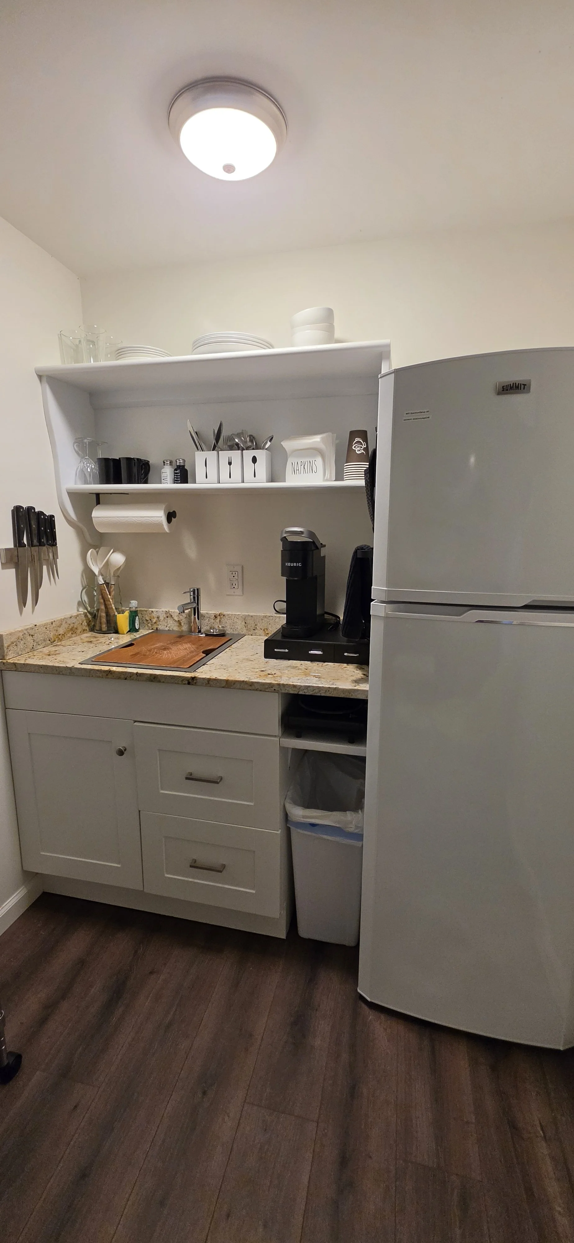 A small kitchen area with a granite countertop, white cabinets, and a white refrigerator. There are kitchen utensils on a magnetic strip, a wooden cutting board, a coffee machine, and shelves with dishes and cups.