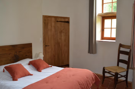 Chambre simple avec un lit en bois, oreillers orange, drap et couverture orange, fenêtre avec vue sur l'extérieur, porte en bois, chaise en bois avec siège en tissu beige, rideau gris.
