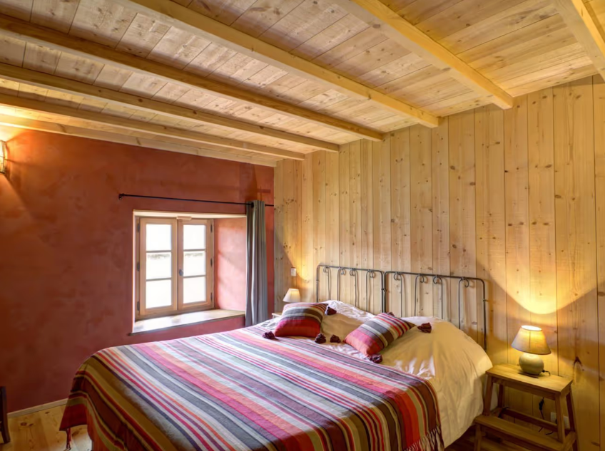 Chambre à coucher avec lit, draps rayés rouges, vert, beige et blanc, fenêtres, murs en bois clair, lampe de chevet, rideaux gris, plafond en bois, ambiance chaleureuse