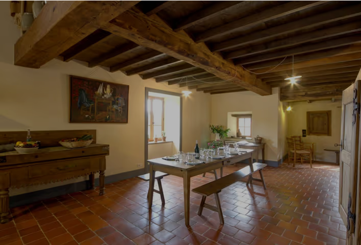 Salle à manger rustique avec table prête pour un repas, sous un plafond en bois avec poutres apparentes, sols en carreaux de terre cuite, murs clairs, fenêtre laissant entrer la lumière naturelle, décorations simples.