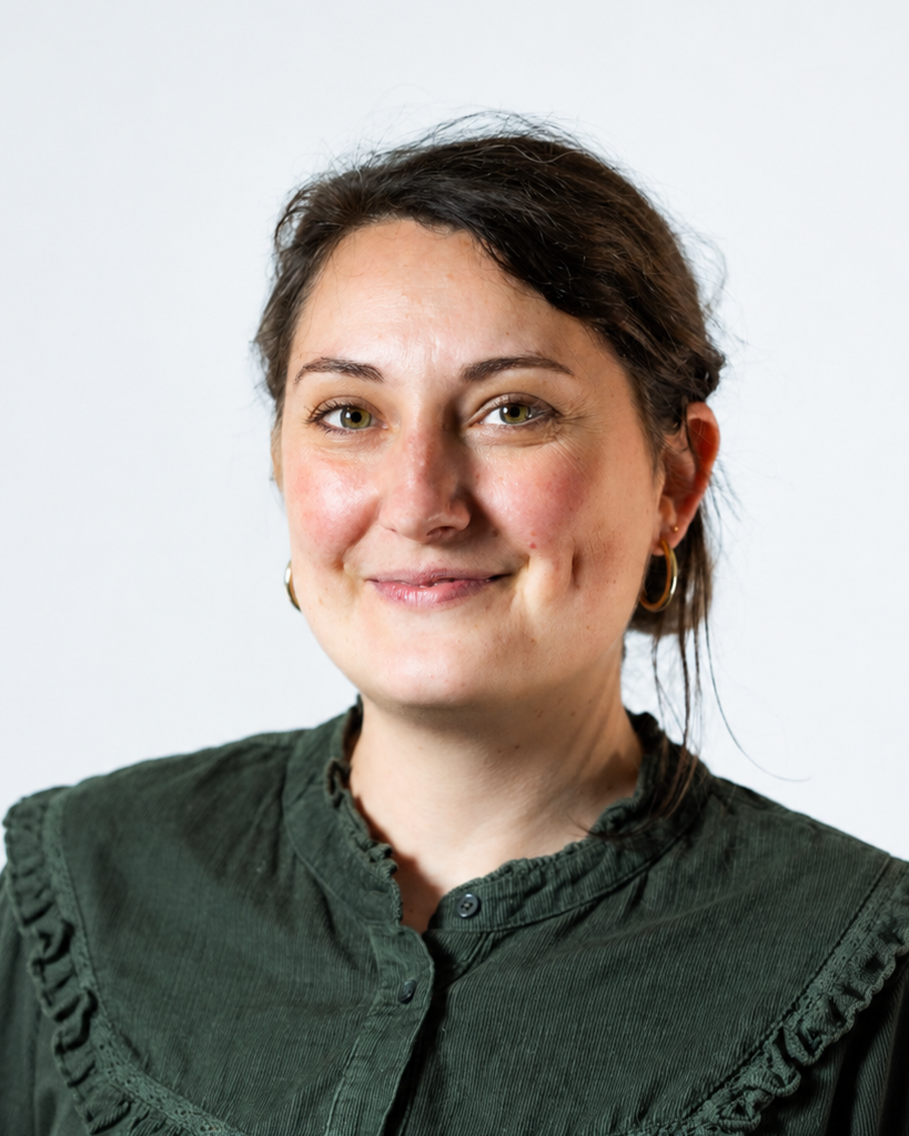 Georgia Blondet - Cheffe cuisinière - Portrait d'une femme souriante, portant une veste verte et des boucles d'oreilles en or, avec un fond blanc.