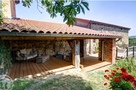 Une véranda en bois avec un toit en tuiles, attachée à une vieille maison en pierre, avec des fauteuils et une table, entourée de plantes et de fleurs.